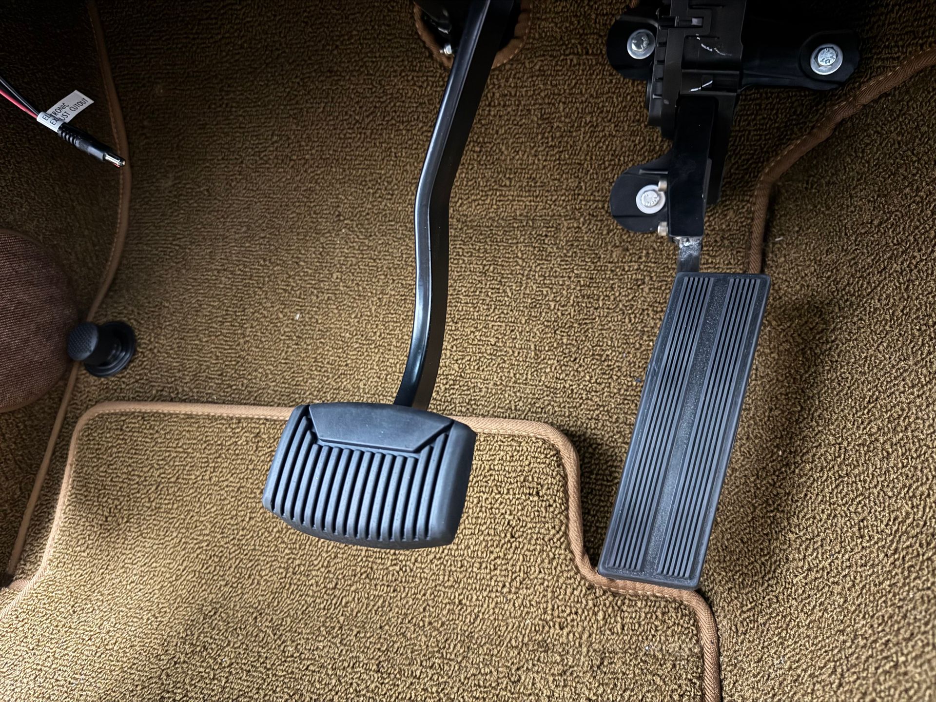 Car pedals under a vehicle dashboard, including a large black brake pedal and a smaller left pedal.