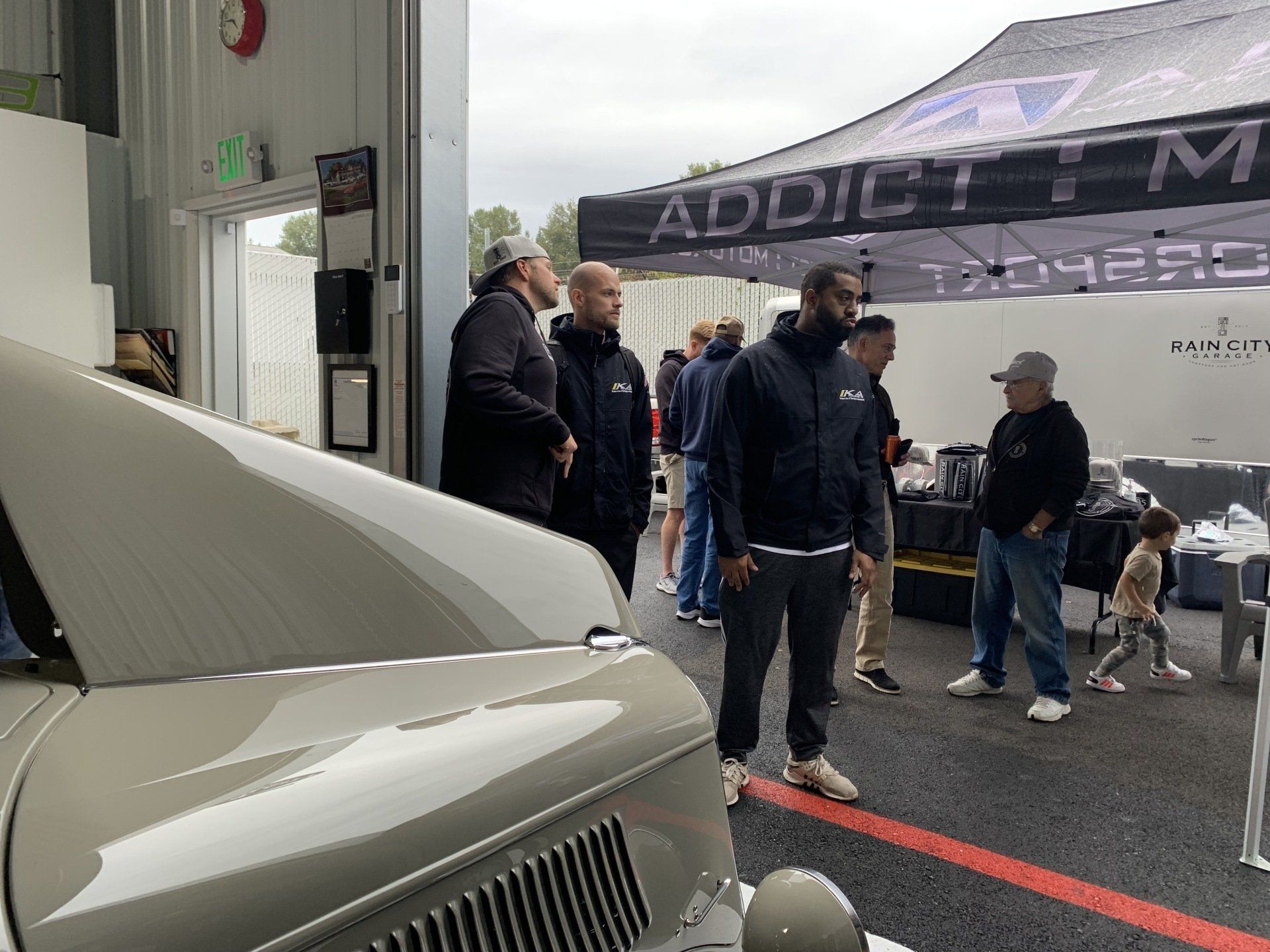 People standing beside a silver car at an outdoor event under a canopy, with a dog nearby.
