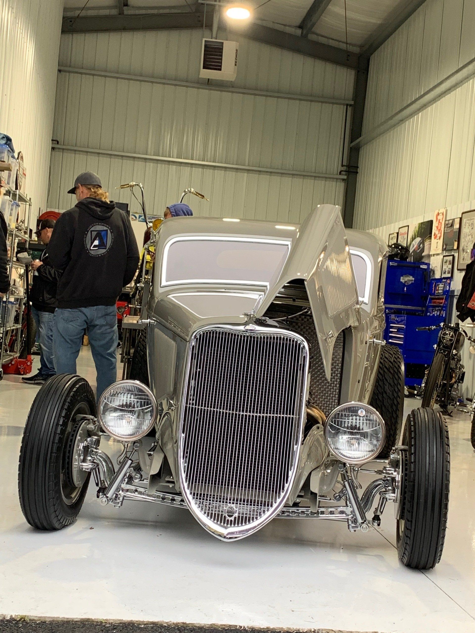Vintage hot rod with exposed engine and chrome grille in a garage, people standing nearby