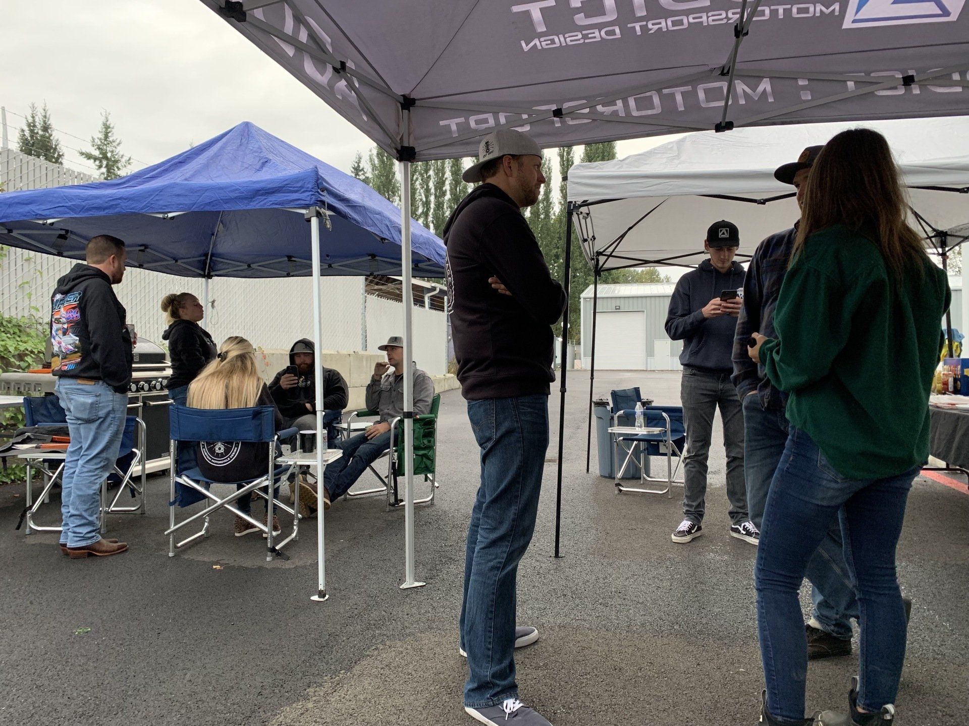 People gathered under blue and gray pop-up tents outdoors on a wet parking lot, chatting and standing around foldable chairs.