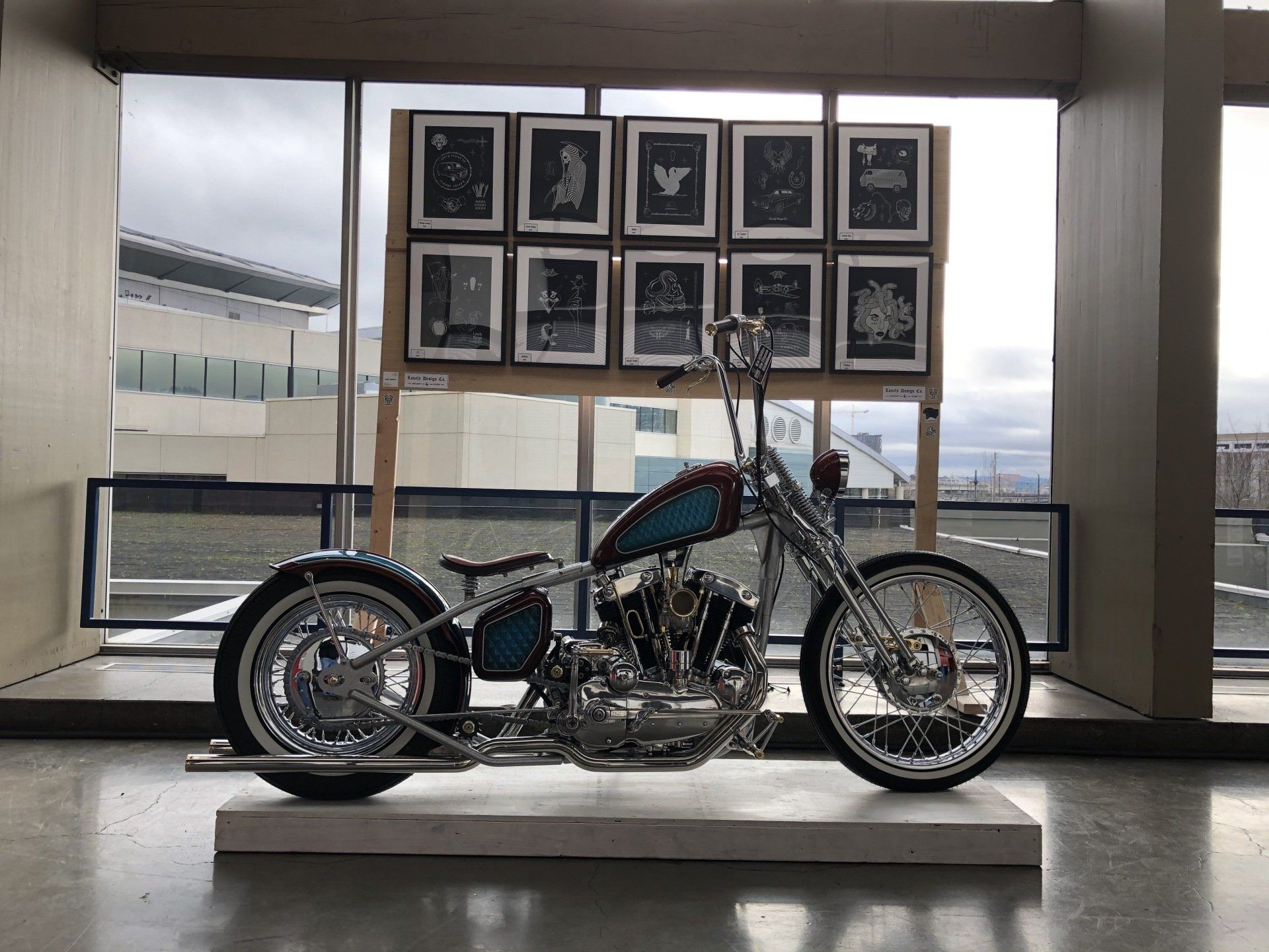 Custom chopper motorcycle displayed indoors on a pedestal in front of a windowed wall with framed photos