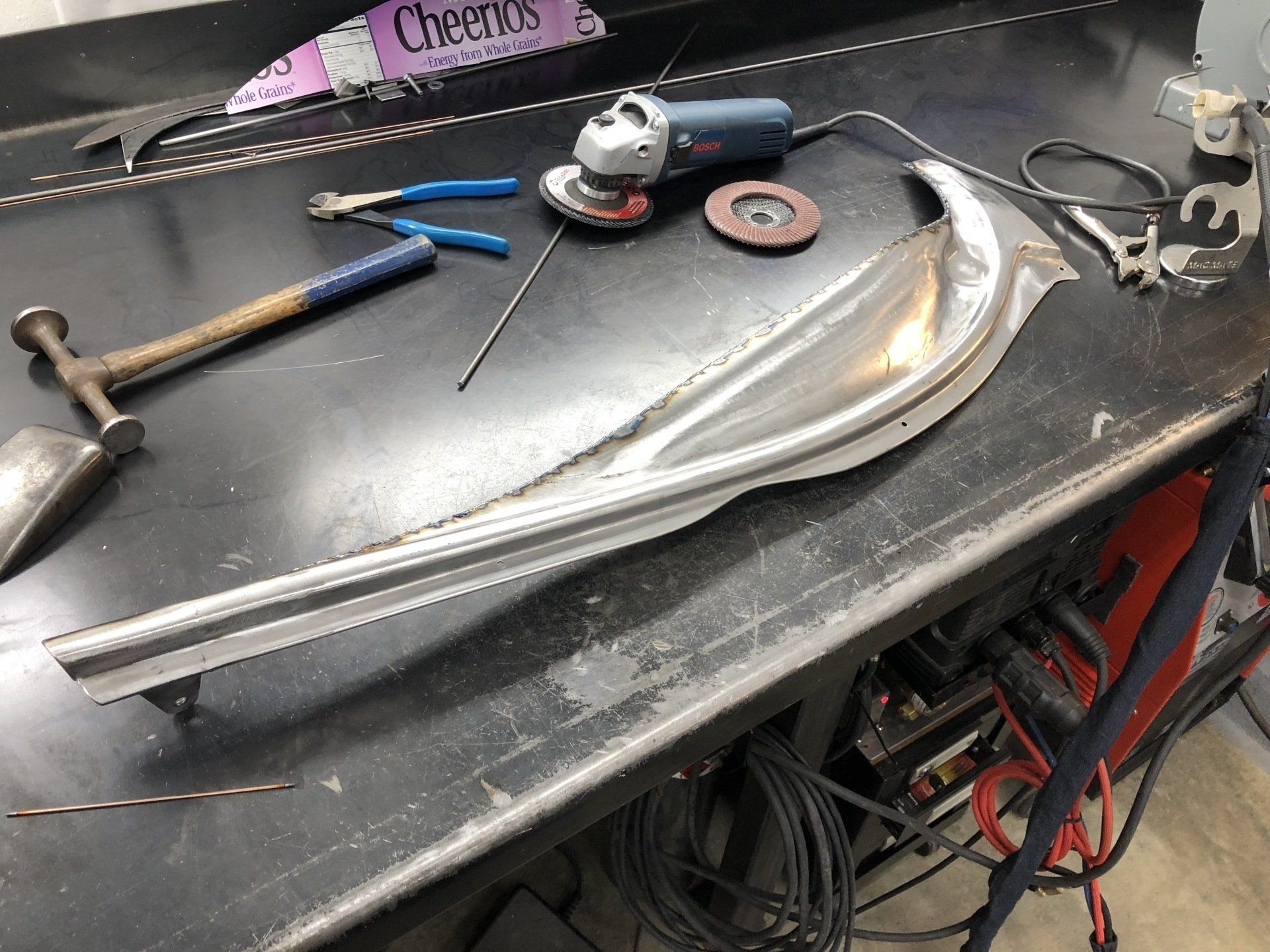 Metal motorcycle body panel on a workbench with tools, including a hammer, grinder, and pliers