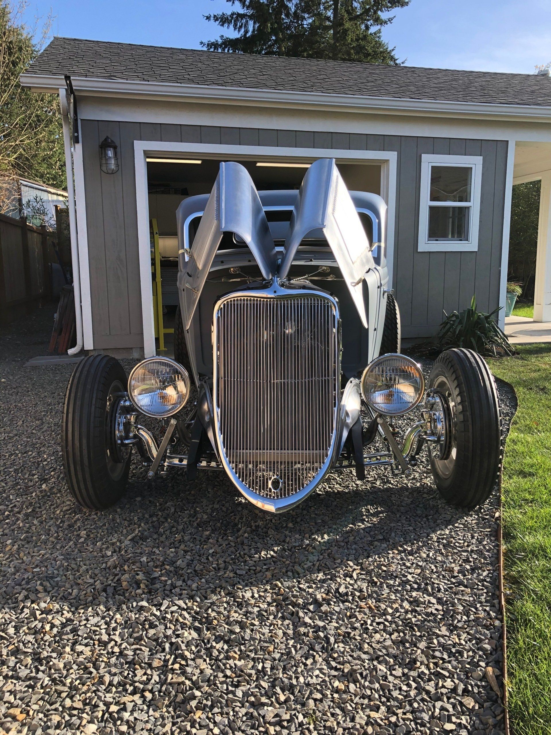 Chrome hot rod parked on gravel in front of a small garage.