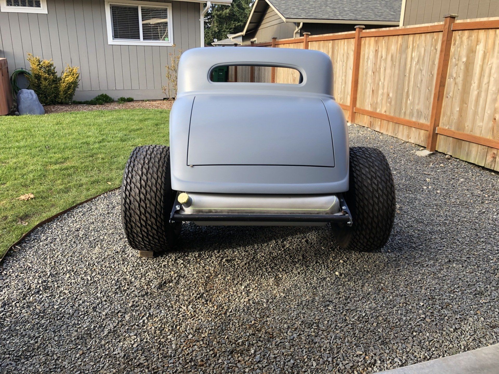 Gray vintage hot rod body on a gravel driveway, viewed from the rear, with a wood fence and house in the background