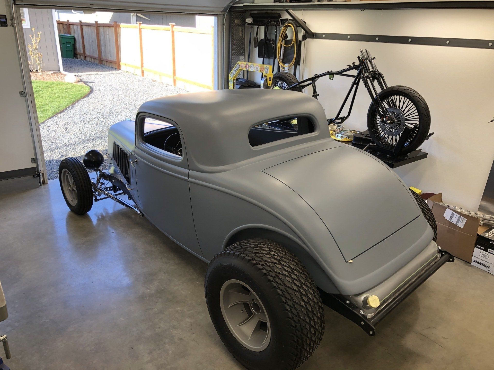 Gray custom hot rod in a garage, with exposed front wheels and a motorcycle in the background.