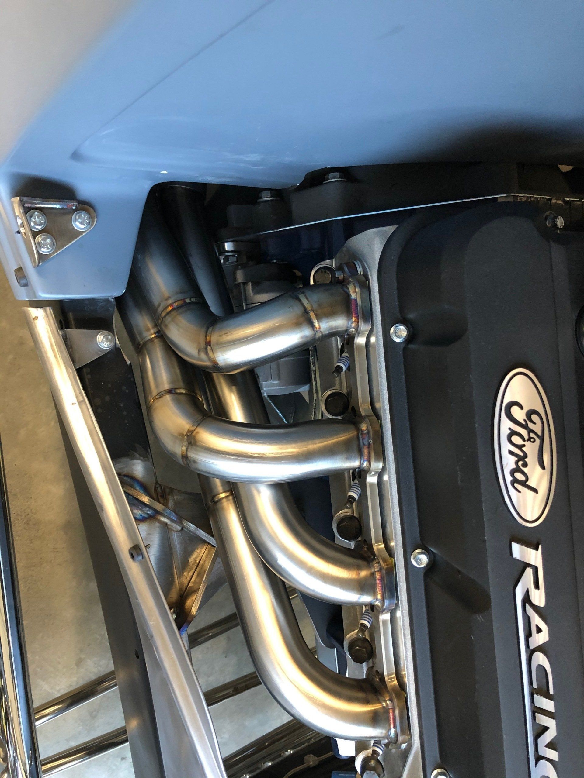 Close-up of a Ford Racing engine bay with polished exhaust headers and black cover under blue bodywork