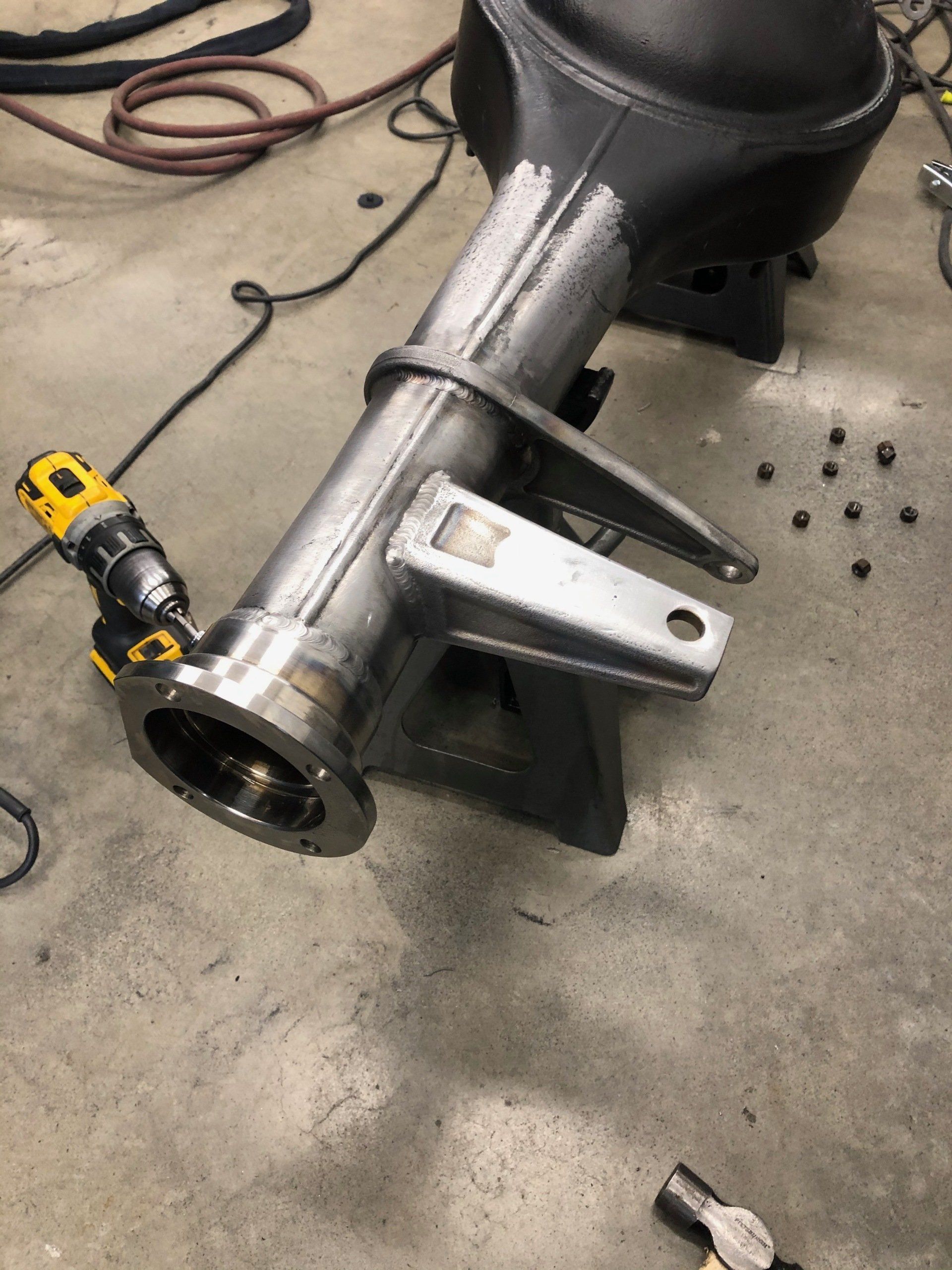 Metal vehicle axle assembly on a concrete floor, with a yellow power tool attached and loose parts nearby.