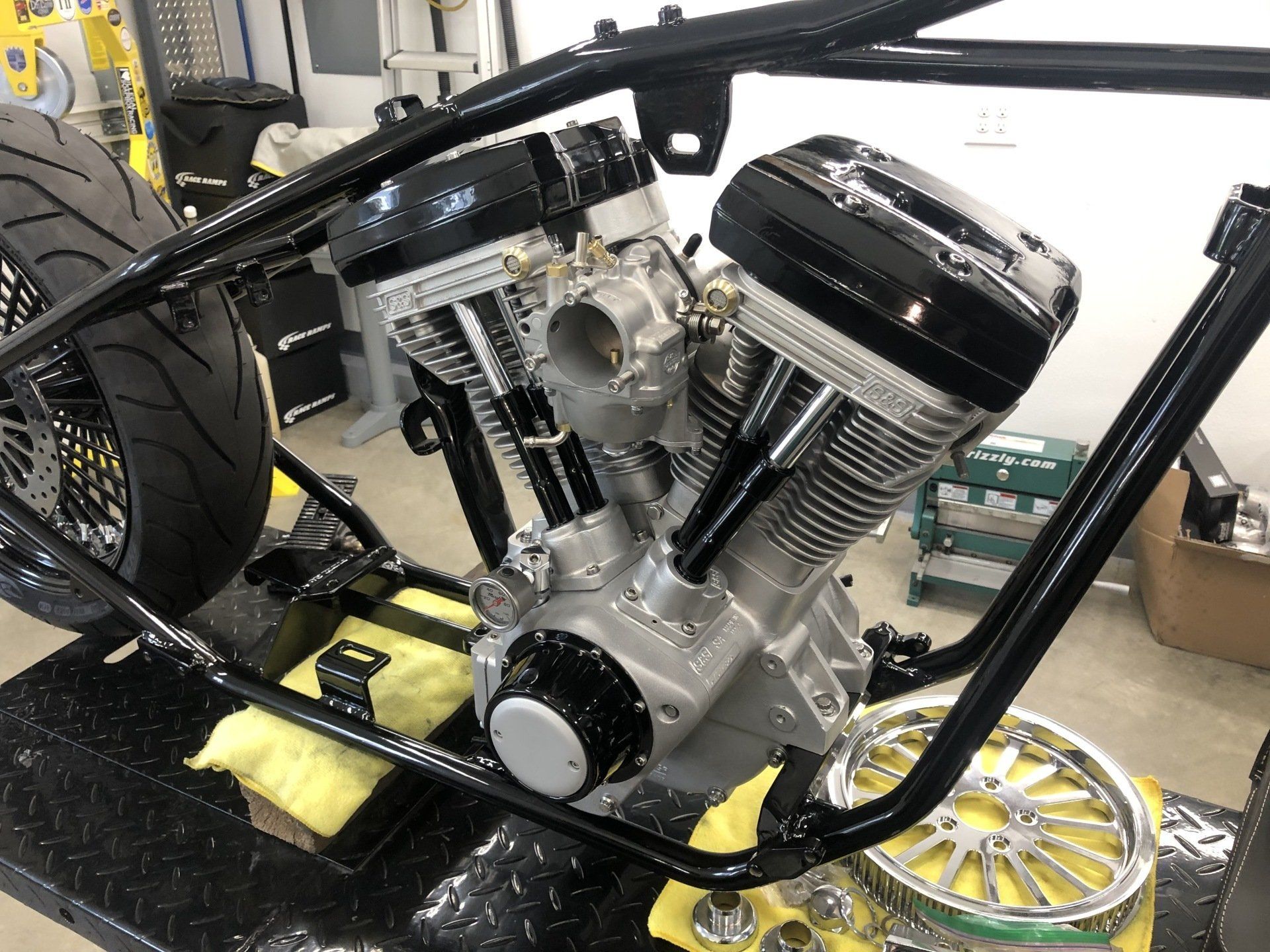 Shiny V-twin motorcycle engine mounted in a black frame, with wheels and workshop tools nearby