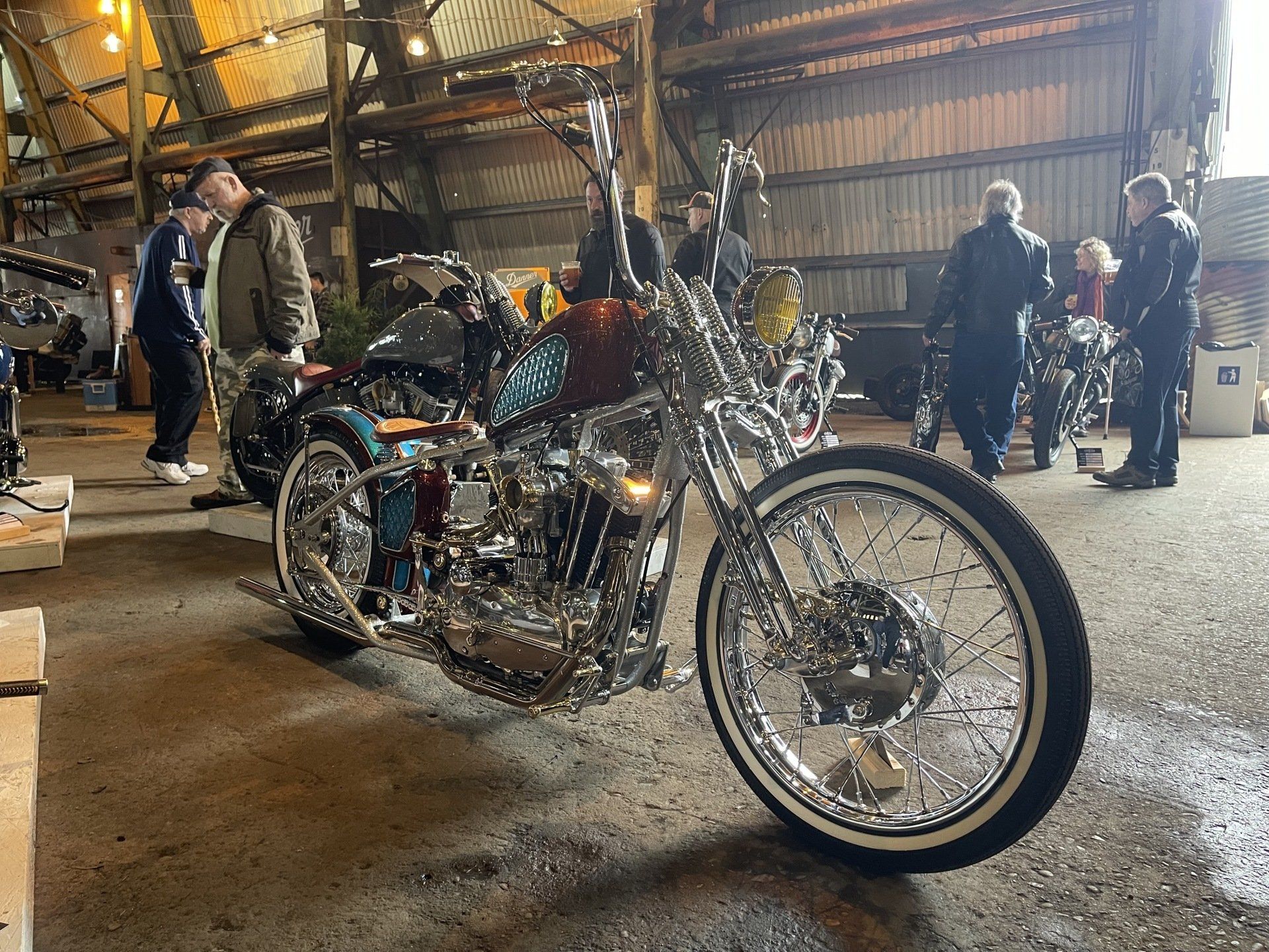 Custom chrome chopper motorcycle indoors at an event, with people in the background.