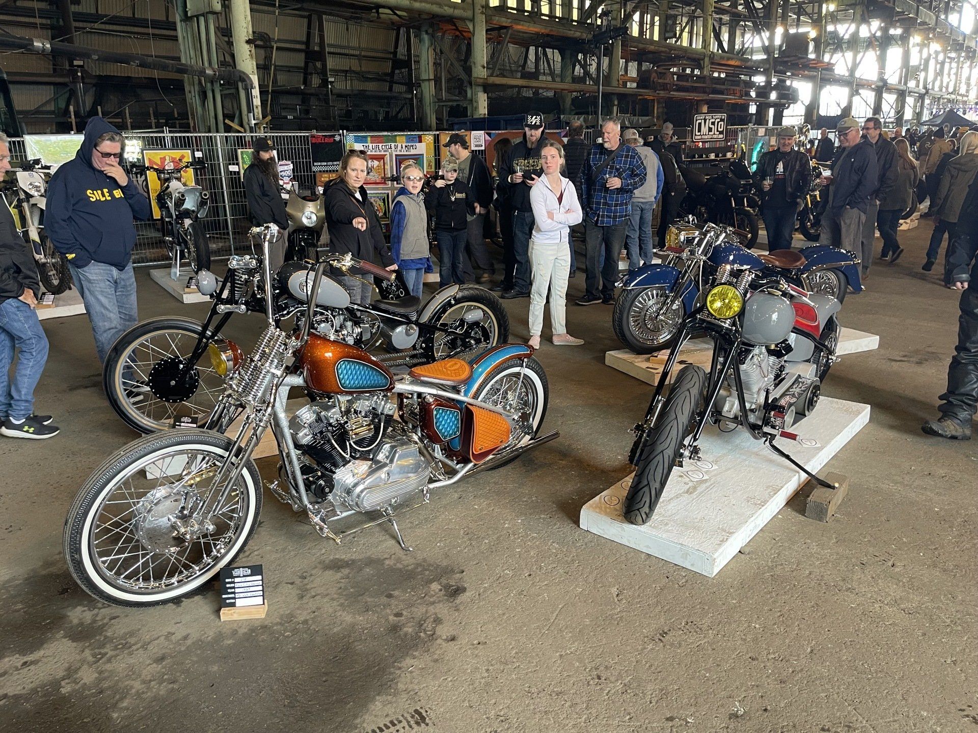Classic custom motorcycles on display at an indoor bike show, with people standing behind them.
