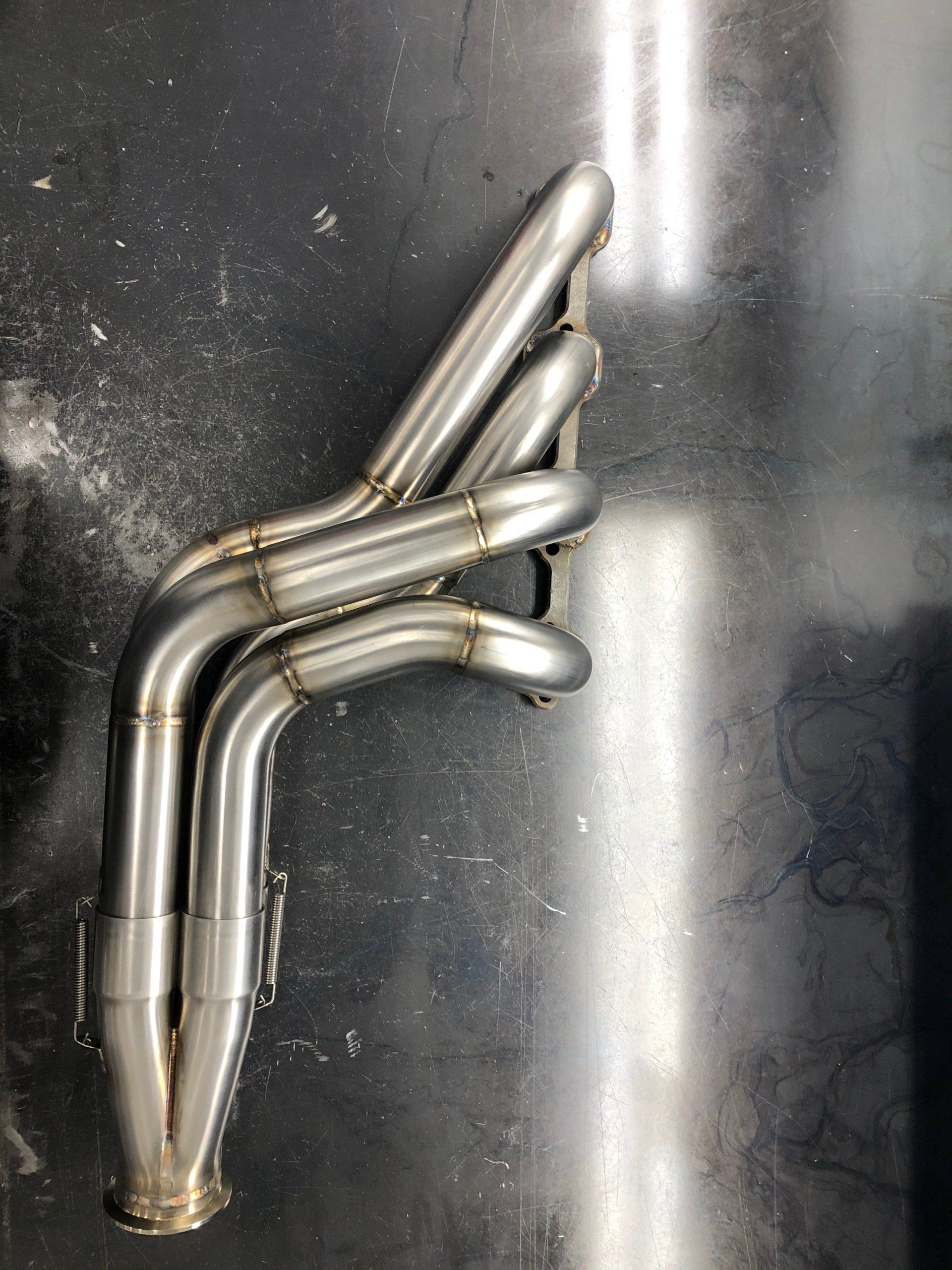 Stainless steel motorcycle exhaust headers on a dark surface, with four curved pipes merging downward.