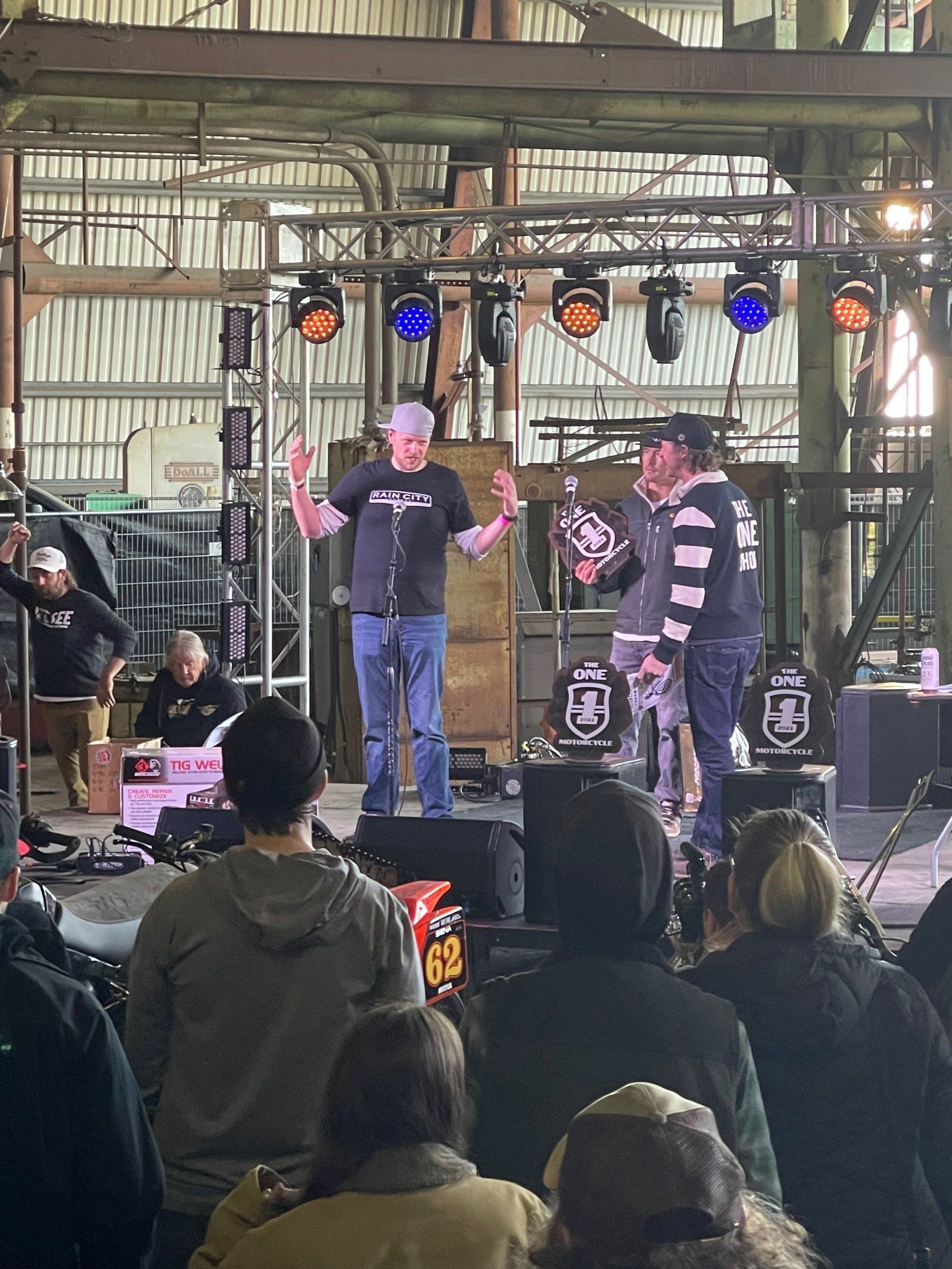 Two men speaking on an indoor stage with colorful lights, seen from an audience in a warehouse venue