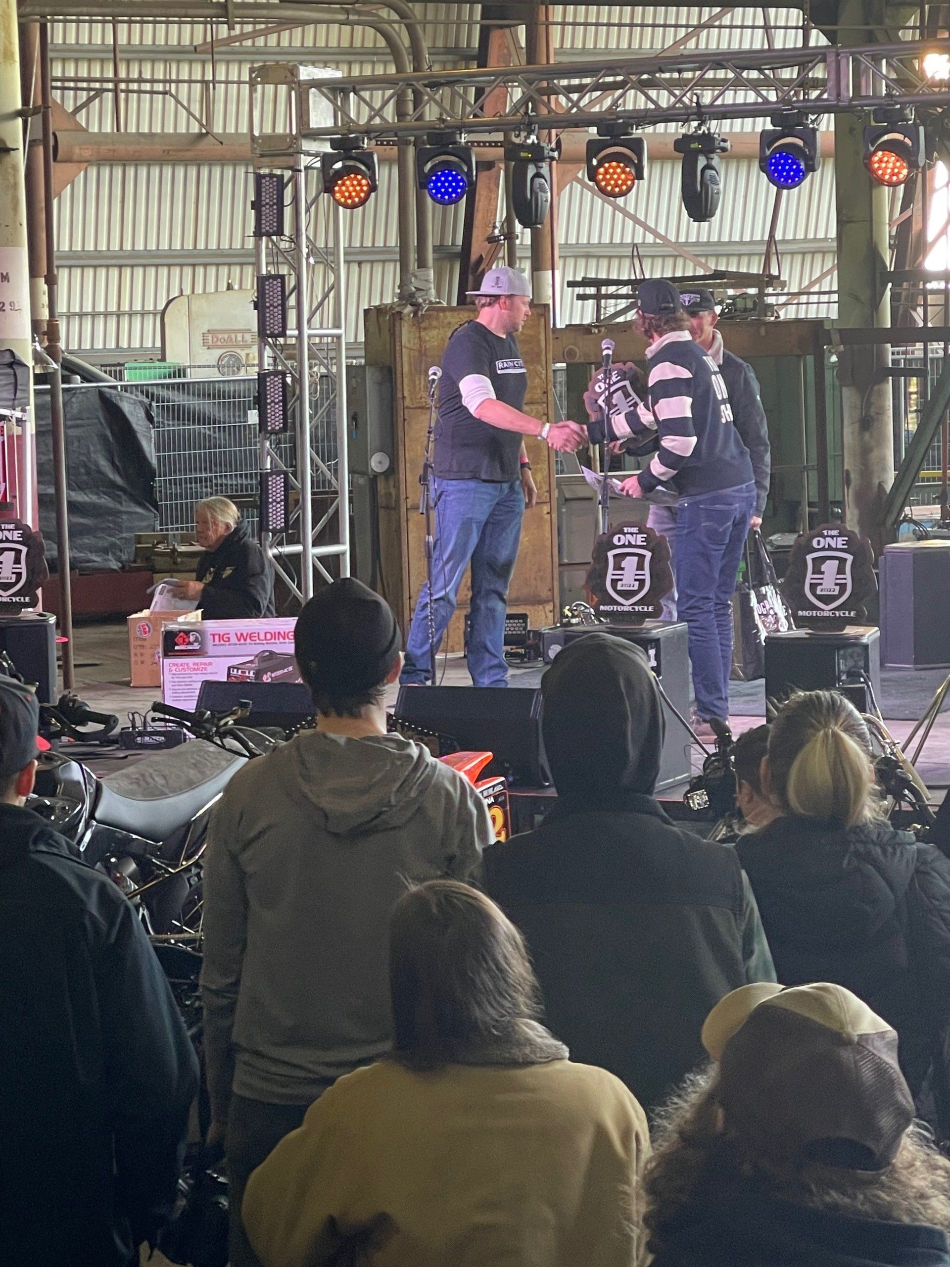 Band on an indoor stage with audience watching, colorful lights and speakers in a warehouse-like venue.