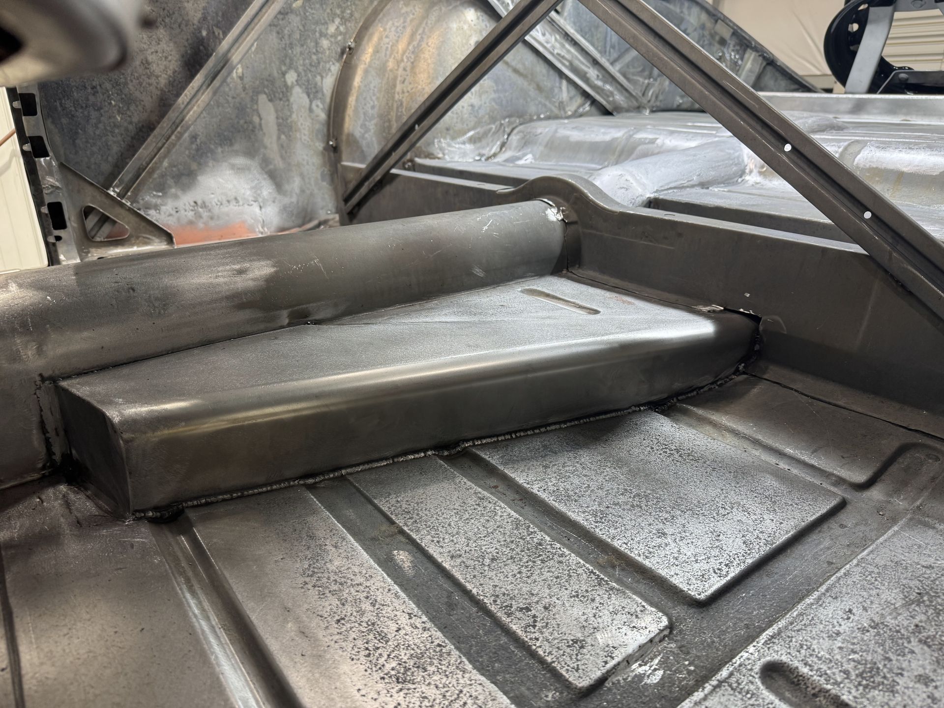 Gray metal industrial duct or chute with sloped panels and a round pipe inside a workshop.