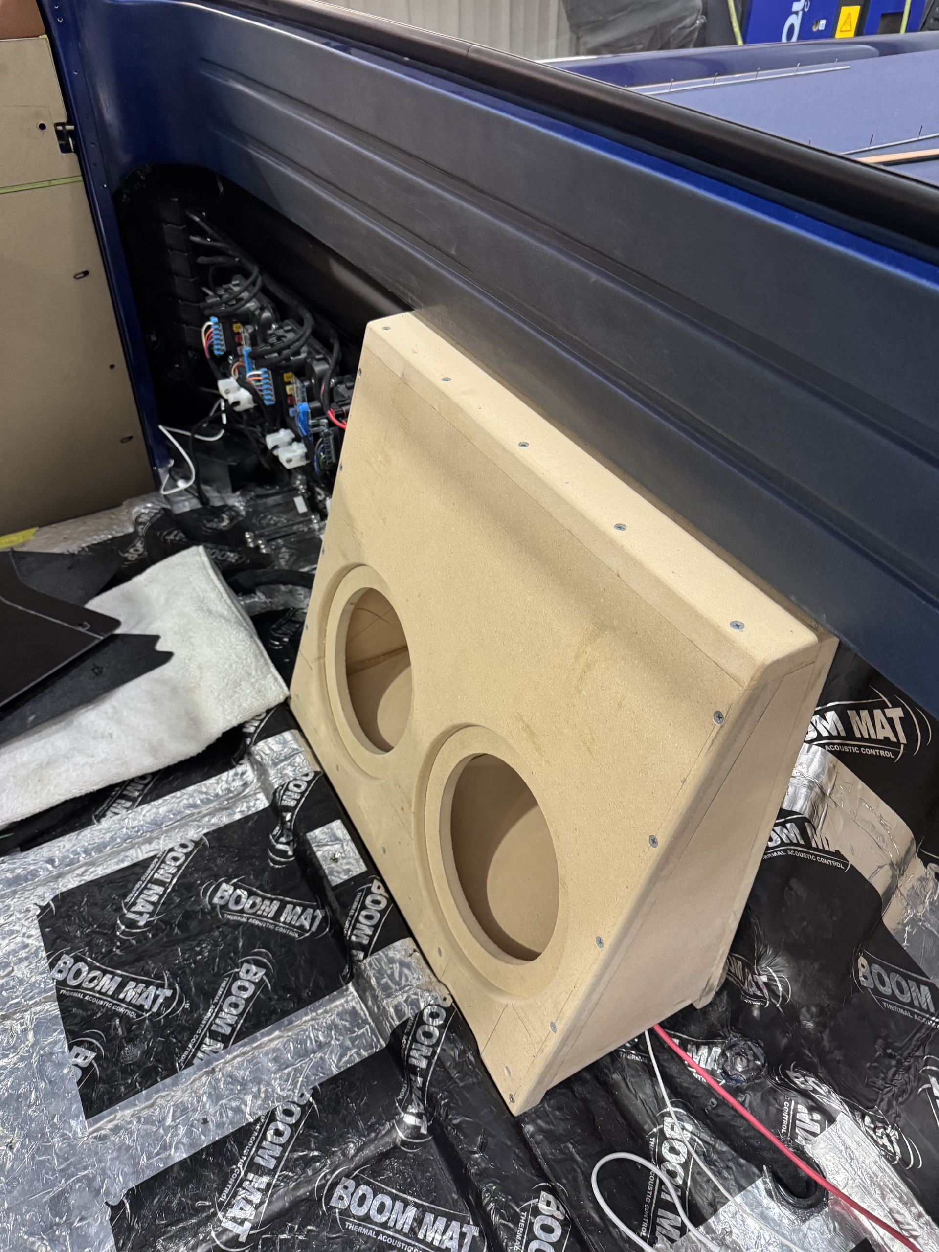 Wooden dual-subwoofer speaker enclosure in the back of a vehicle, beside a black storage bin.