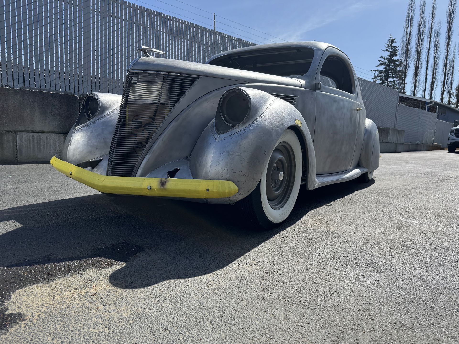 Vintage gray custom car with yellow front bumper parked on pavement near a fence