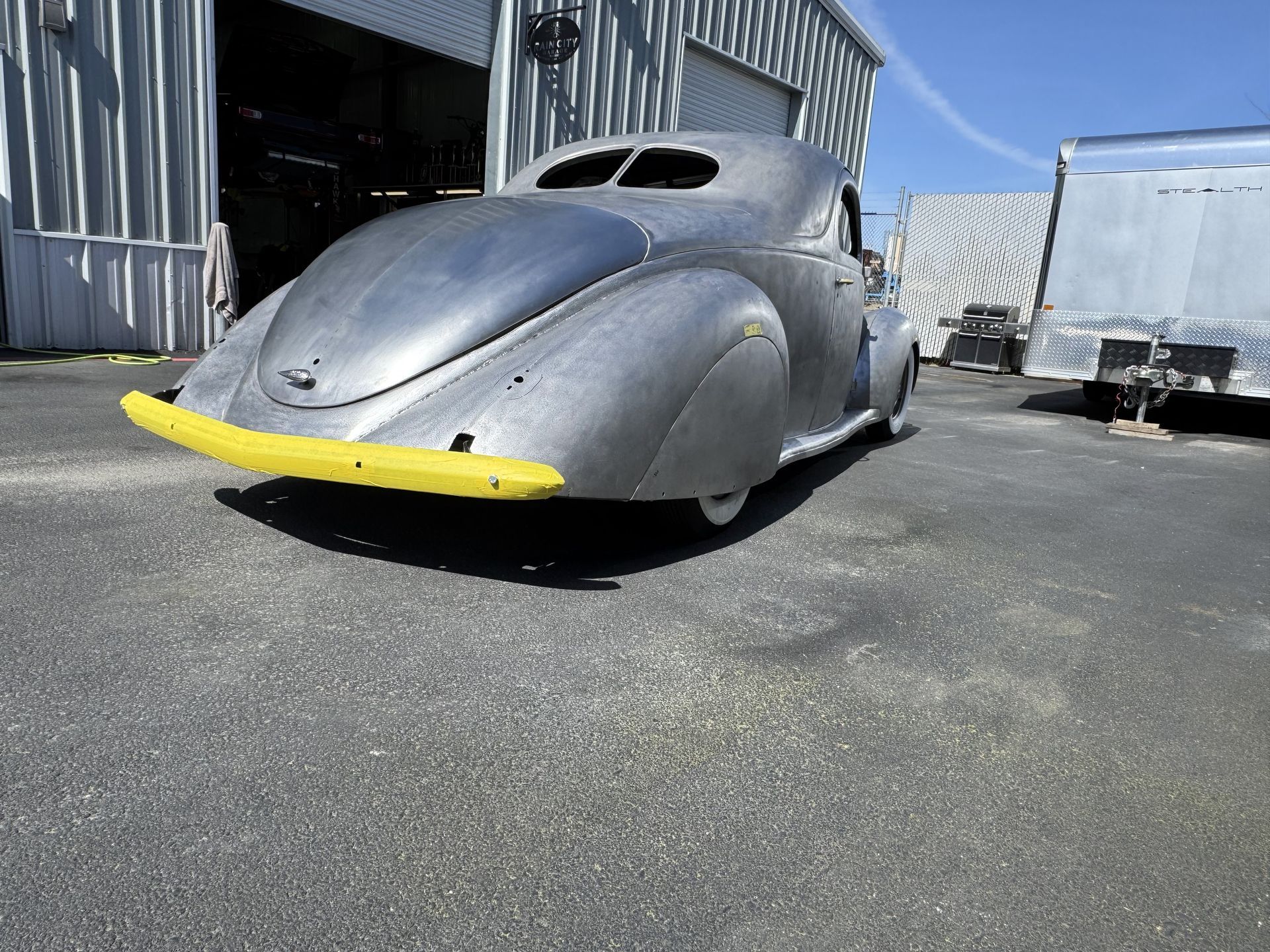 Silver vintage car with a yellow front bumper parked beside a warehouse