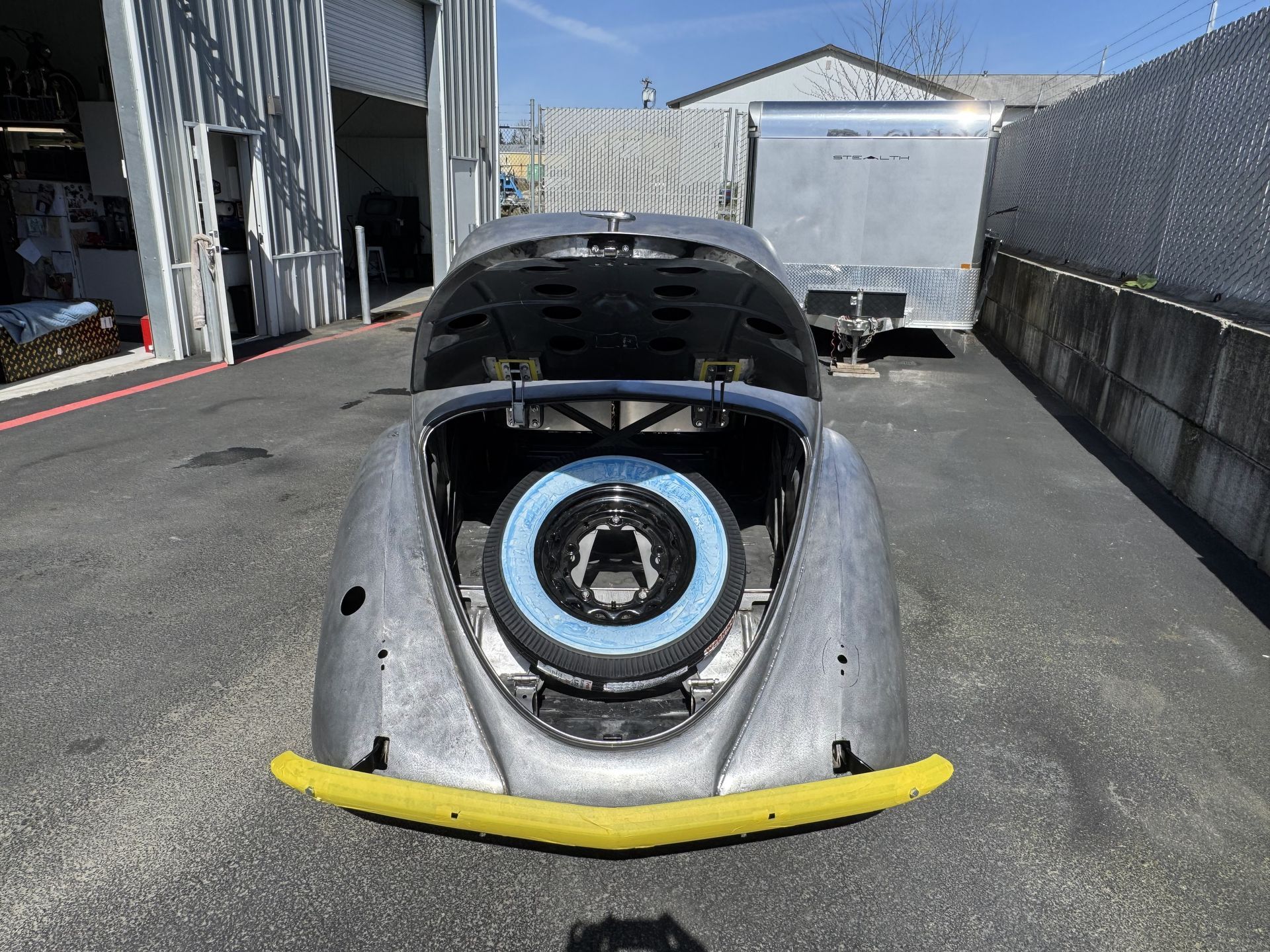 Rear view of a silver VW Beetle with its engine exposed, parked in a lot under bright sunlight.