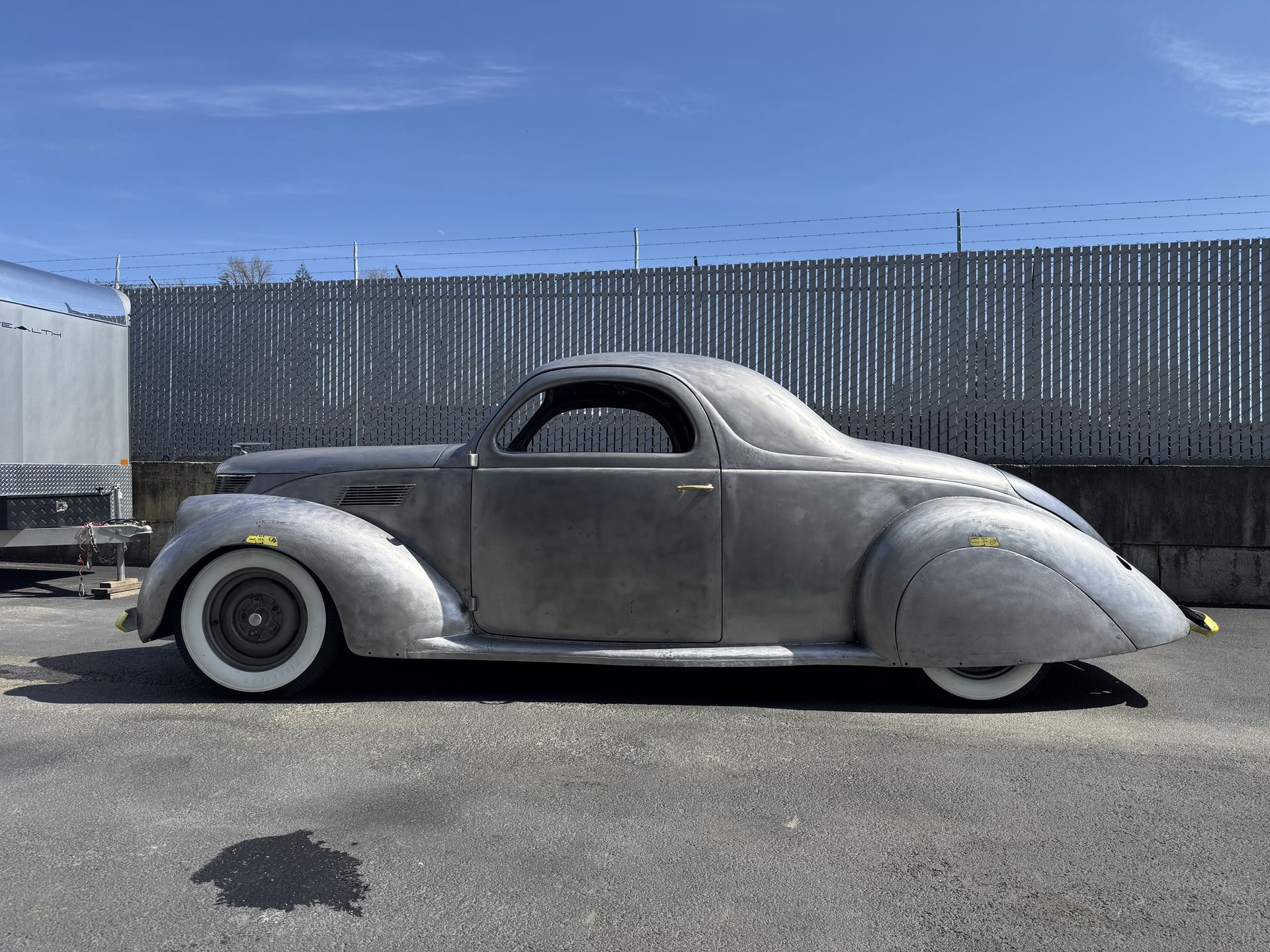 Vintage gray coupe parked outdoors beside a truck and fence.