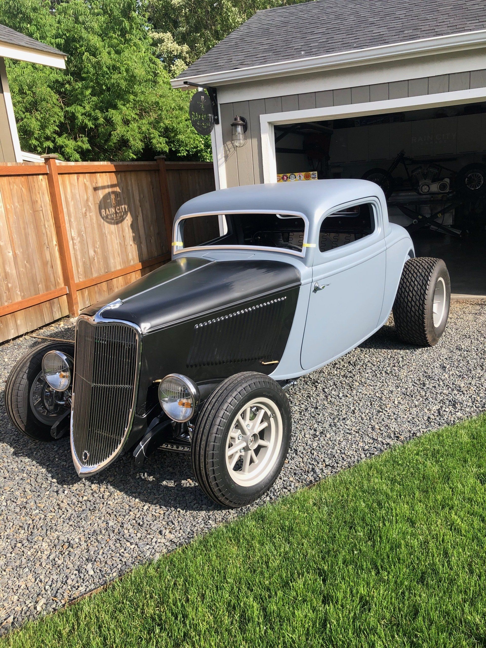 Classic black-and-gray hot rod parked on a gravel driveway outside a garage.