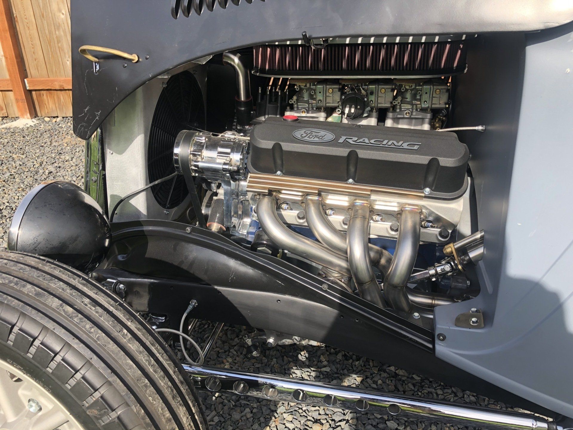 Classic car engine bay with chrome headers and black engine components under a gray hood.