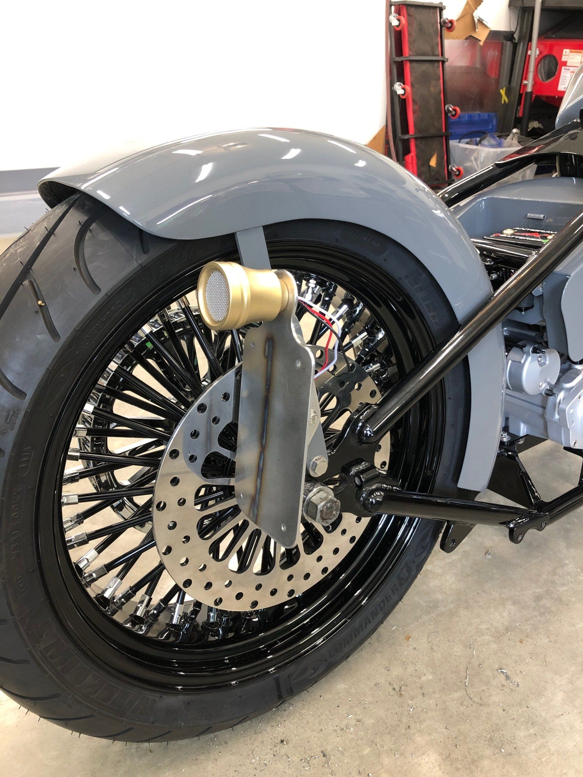 Custom motorcycle wheel and fender in a workshop, with a black tire and chrome exhaust.
