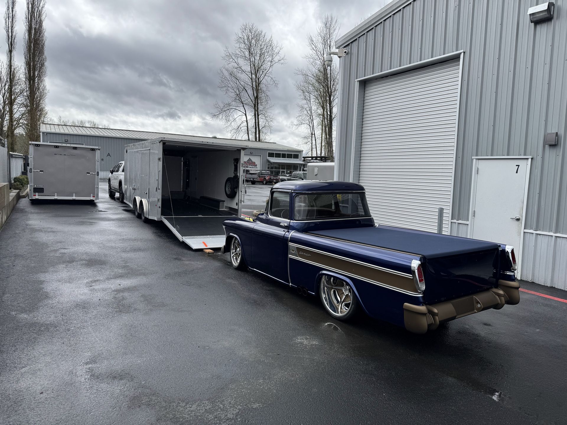 Blue classic pickup truck with an open trailer parked on a wet industrial lot beside a warehouse