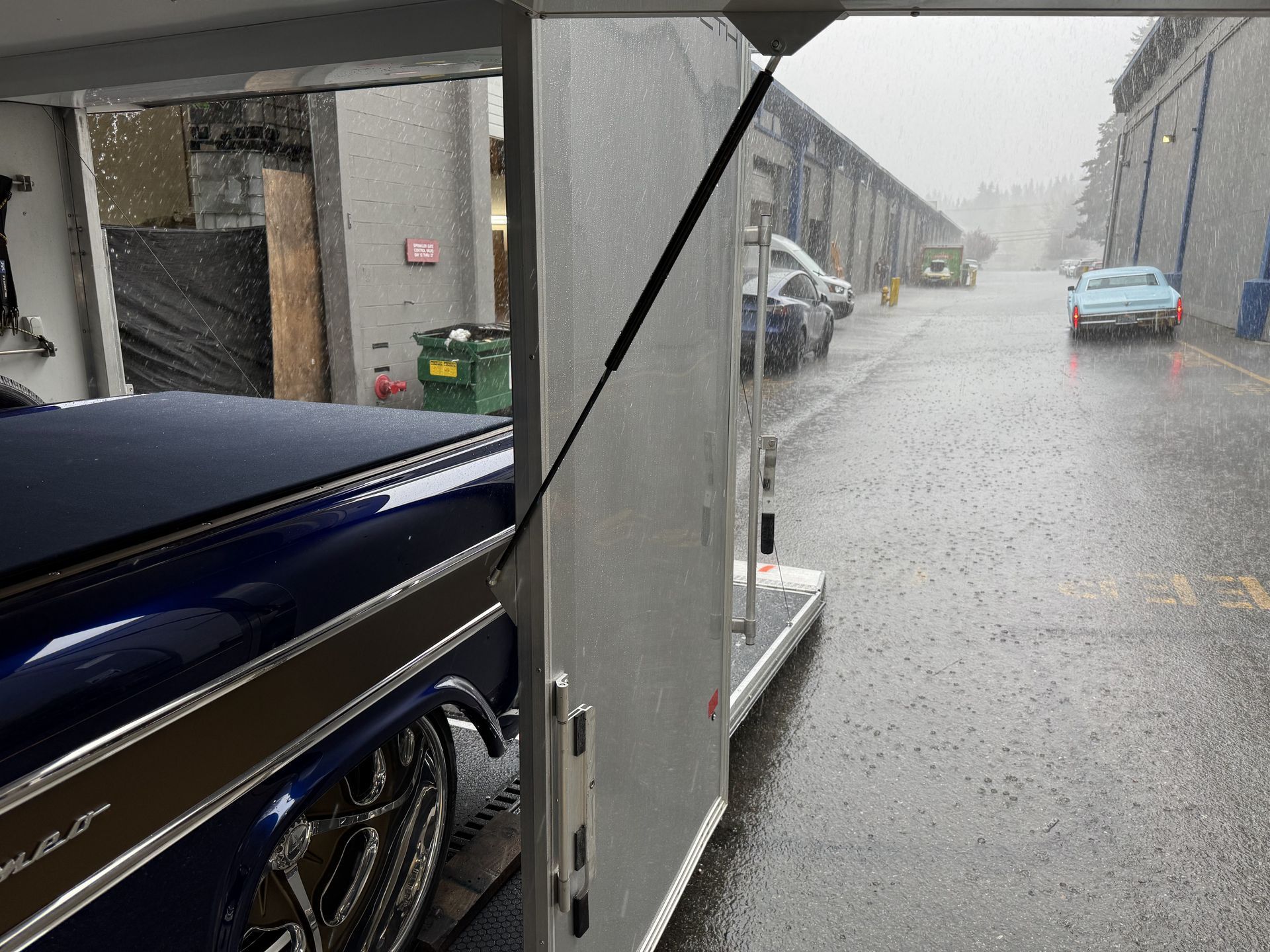 Rainy street view from a carport, with a blue car in the foreground and parked cars along a wet lane.