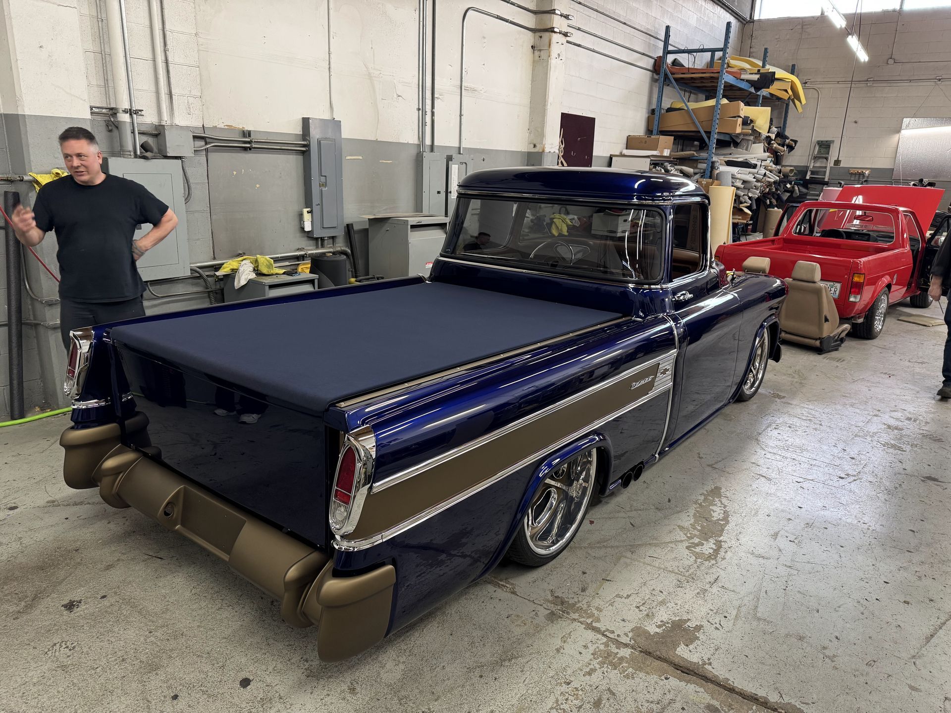 Custom dark-blue classic pickup truck in a garage, with a red car behind it and a person standing nearby.