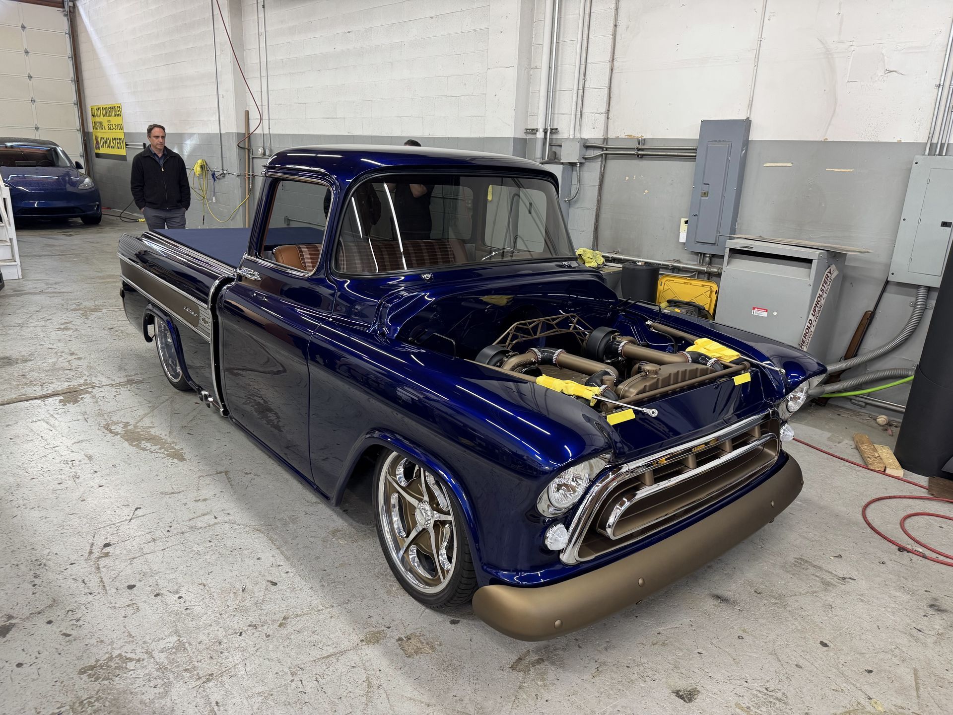 Blue classic pickup truck in a garage, hood open, showing engine; a person stands in the background.