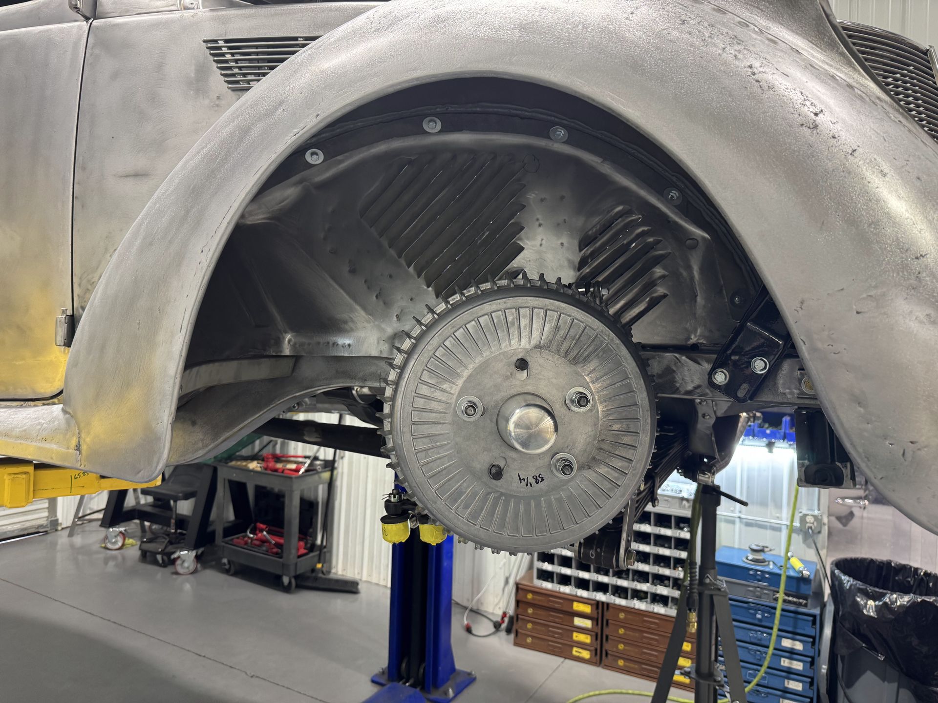 Car raised in a garage, exposing a large circular wheel and undercarriage with tools in the background