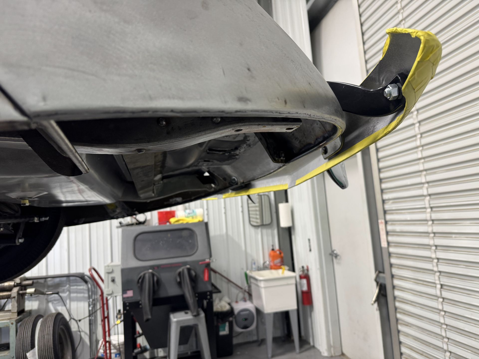 Underside of a car on a lift in an auto shop, with a yellow strap and garage equipment nearby