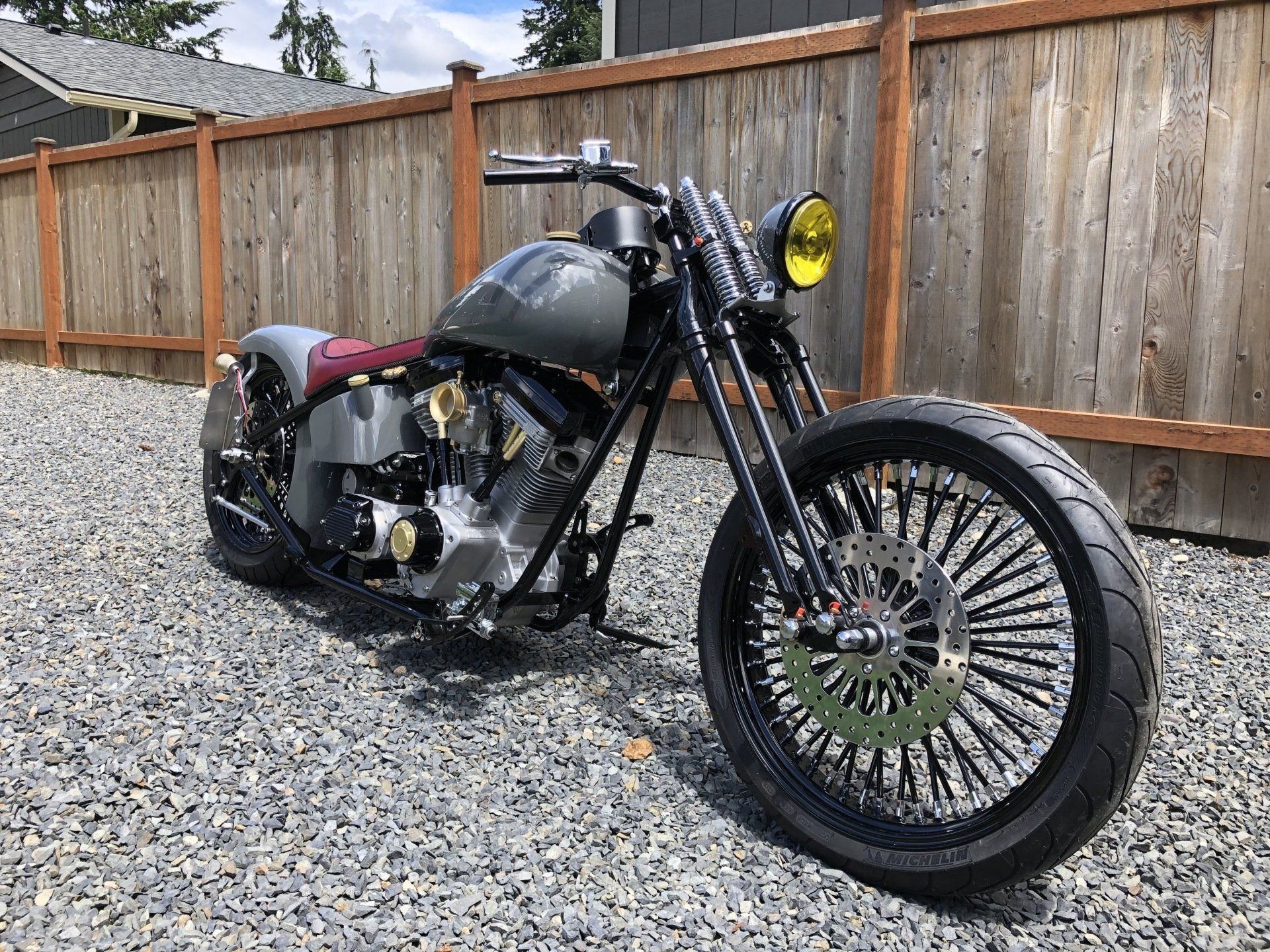Custom black motorcycle with yellow headlight parked on gravel beside a wooden fence.