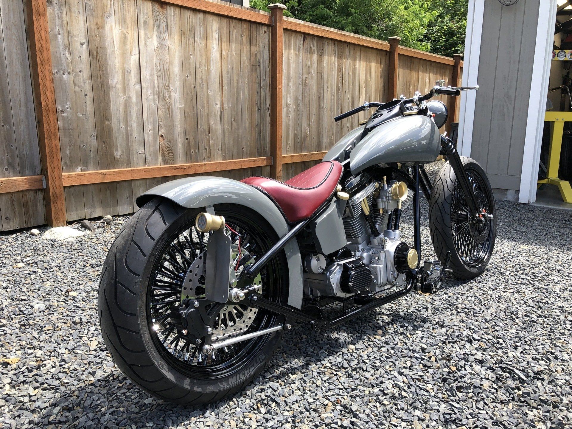 Custom silver-and-black motorcycle with a maroon seat parked on gravel beside a wooden fence.