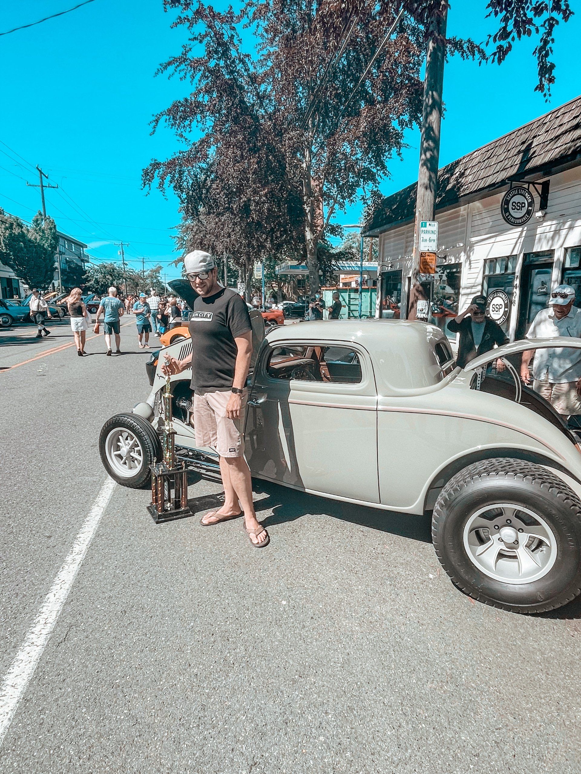 Man standing beside a gray vintage hot rod on a sunny street during a car show