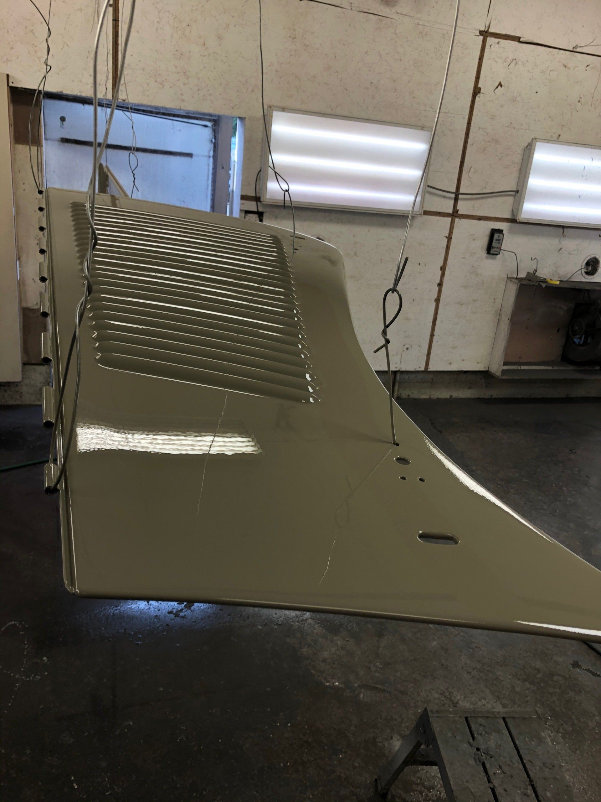 Olive-green metal automotive panel hanging in a spray booth, with perforated section and overhead lights