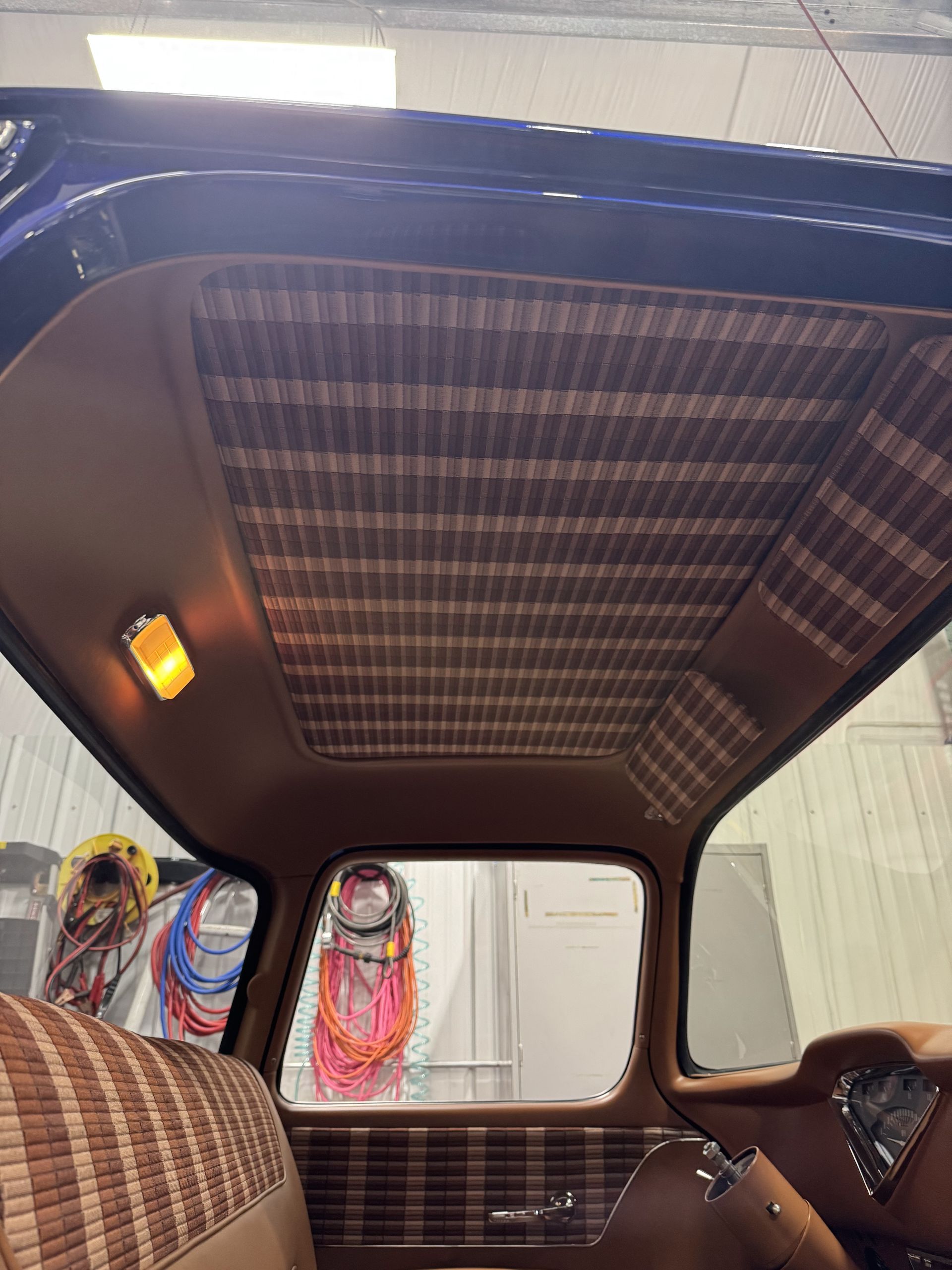 Vintage car interior with plaid seats, patterned roof lining, and coiled cables visible in the back.