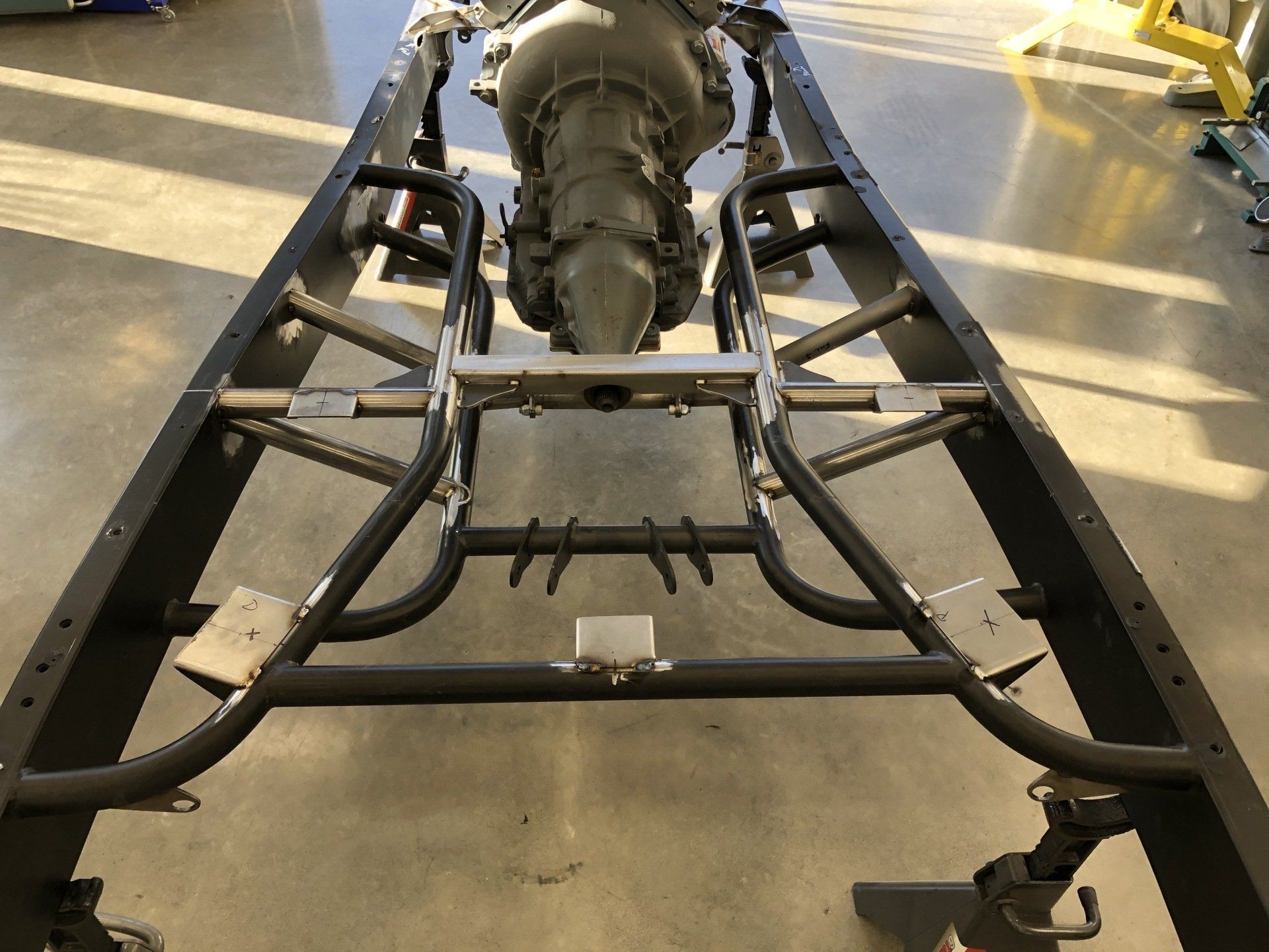 Race car chassis frame on a workshop floor, viewed from the front with suspension arms visible