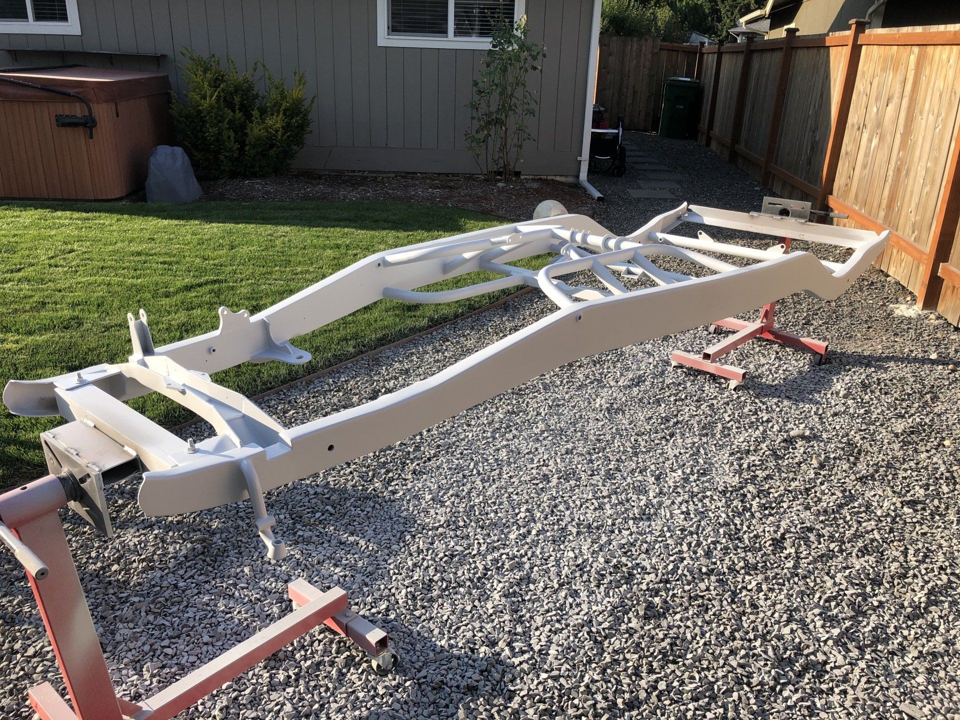 White utility trailer chassis on gravel in a driveway, with red supports and a wooden fence nearby