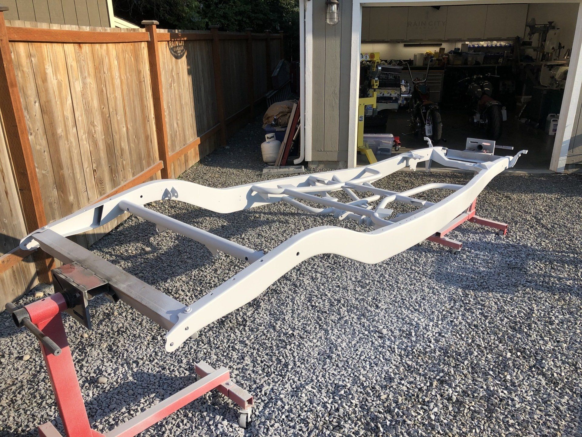 White trailer frame on gravel drive outside an open garage, with a red jack stand nearby.