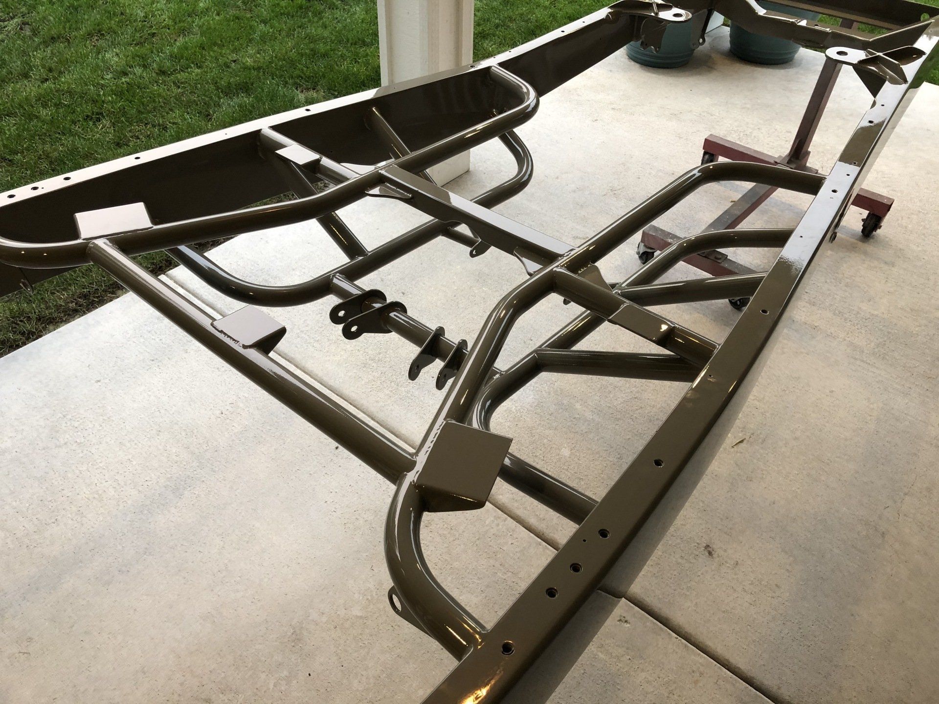 Black metal trailer chassis frame on a concrete porch, with wheels and curved support bars attached.
