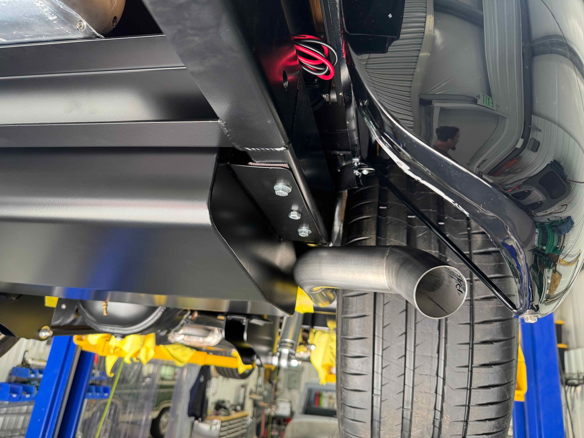 Undercarriage of a car on a lift, showing rear tire, suspension, and exhaust with exposed wiring.