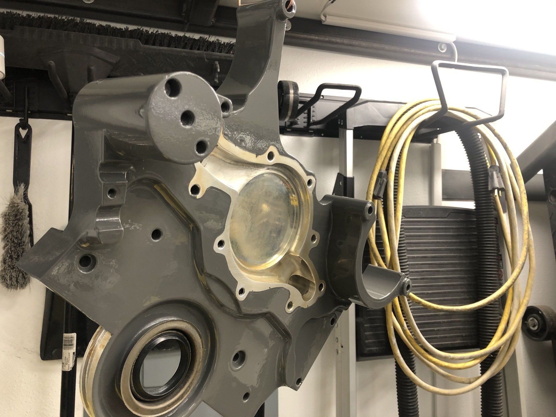 Gray engine block hanging in a workshop, with yellow coiled hose and metal piping nearby.