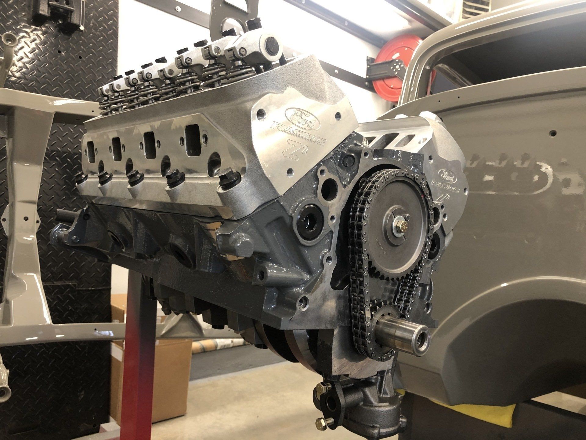 V8 engine mounted in a workshop, with exposed timing chain and silver cylinder heads.