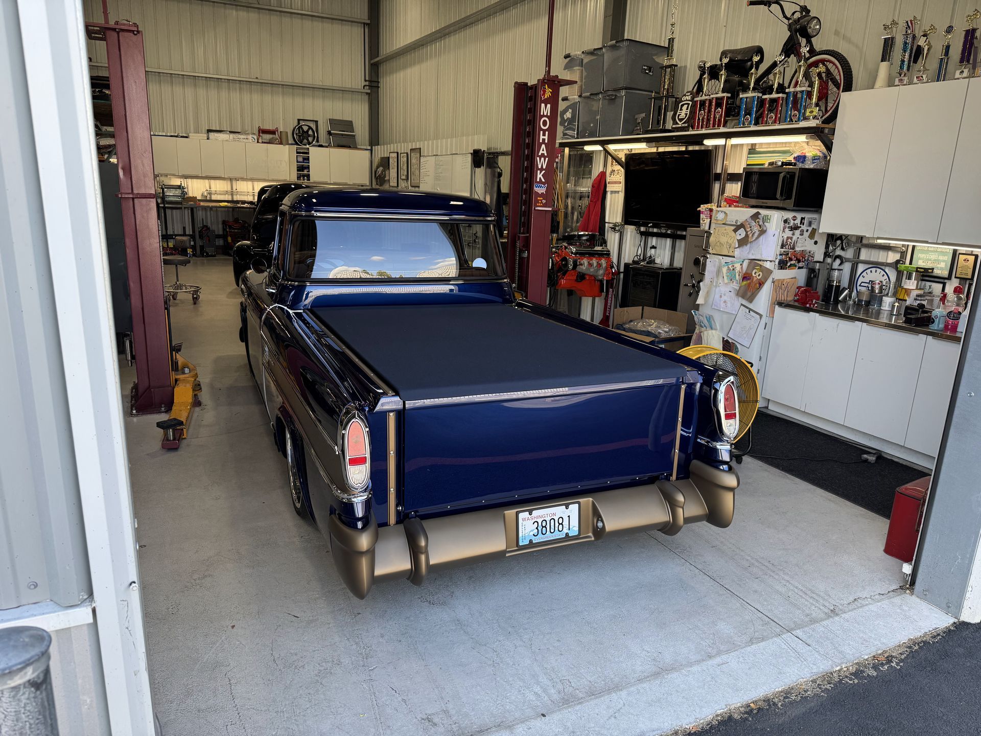Dark blue classic car parked in a garage with tools and cabinets around it