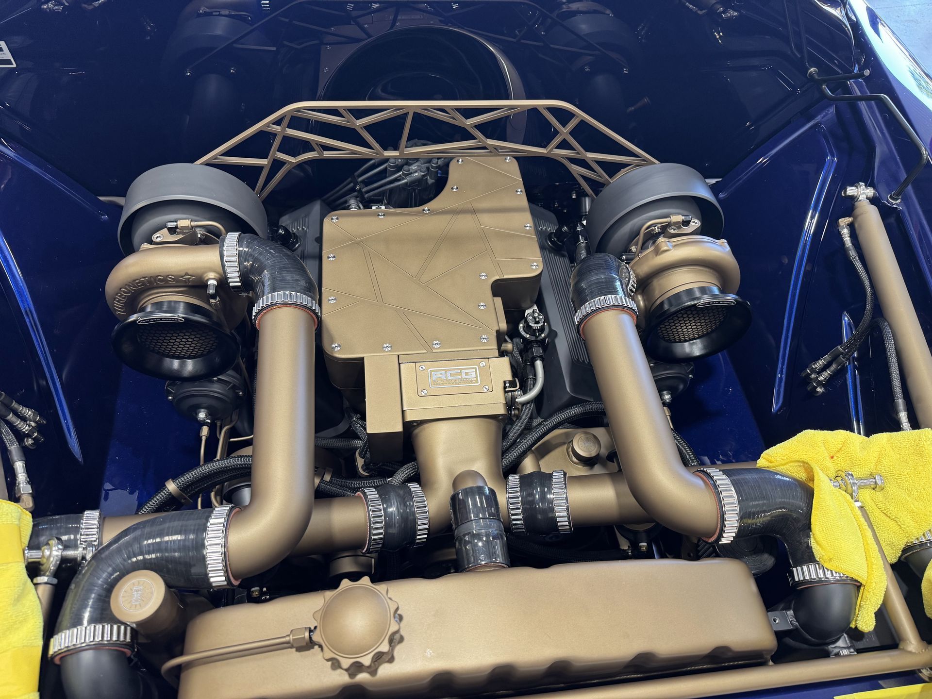 Close-up of a car’s rear engine bay with twin turbos and tan piping against a blue body.