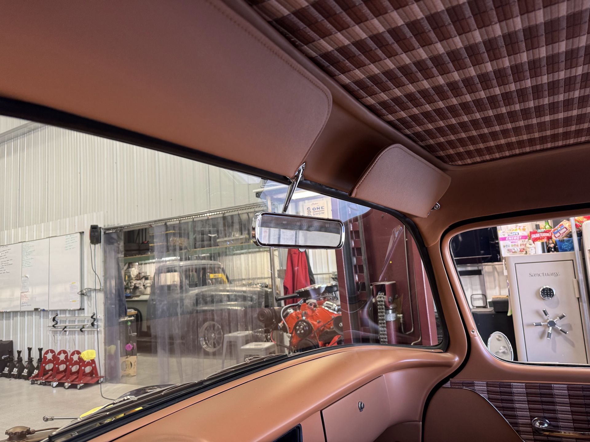 Inside a vintage car, looking out through the rear window at a workshop with equipment and a vehicle outside