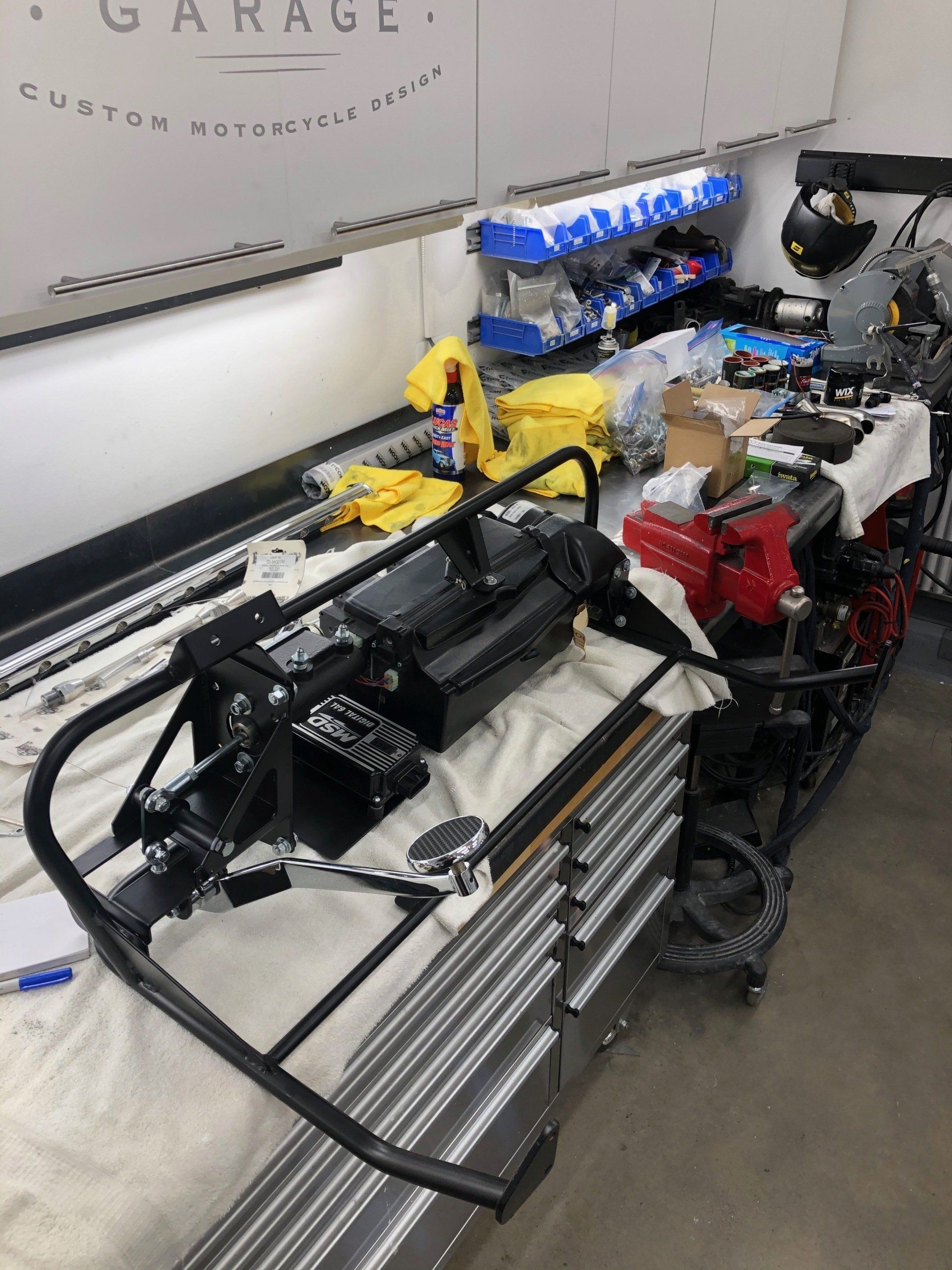 A cluttered garage workbench with a black frame, tools, and colorful mechanical parts under bright lights