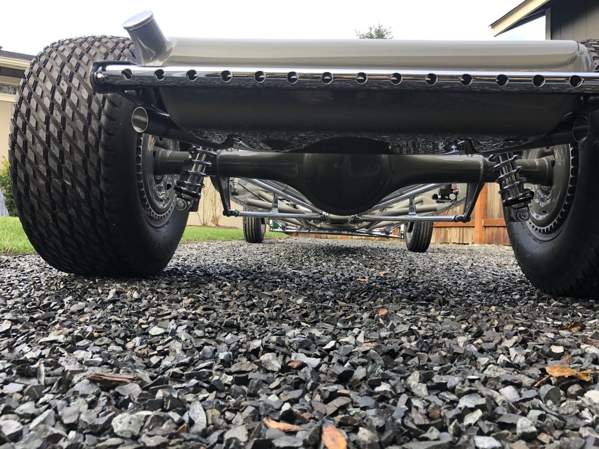 Low-angle view of a vehicle’s undercarriage and large tires on gravel, with a chrome exhaust pipe visible