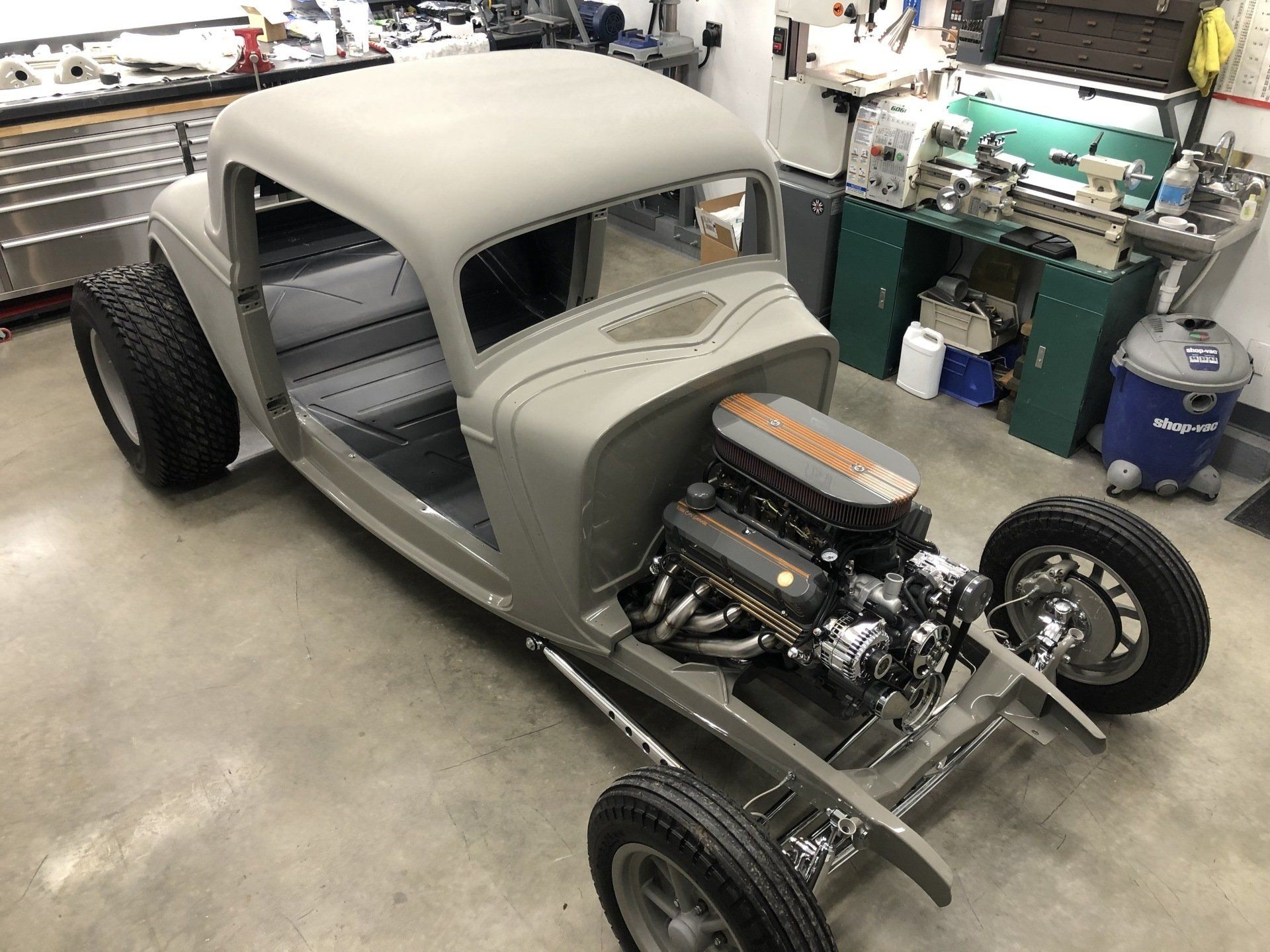 Gray custom hot rod chassis in a workshop, front-engine with exposed wheels and body shell.